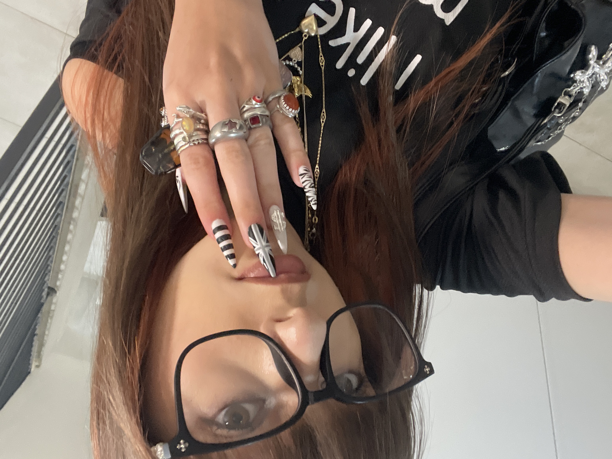 The image is an upside-down selfie of a person with long, brown hair, wearing glasses with black frames. The person's hand is prominently displayed in the foreground, adorned with multiple rings and long, decorated fingernails. A black t-shirt and a black bag with a silver chain are visible in the background. The specific location in Raanana, Israel cannot be determined from the image alone.