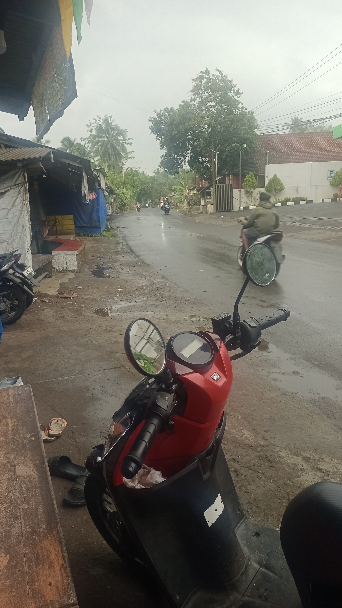An overcast, cloudy day is depicted along a wet, asphalt road in Cikalong, Indonesia. In the foreground, a red and black scooter is parked on a dirt and gravel shoulder. Its handlebars are visible, along with a large, round rearview mirror reflecting trees. To the left of the scooter, a weathered wooden table is partially in frame, with a pair of black sandals and a single flip-flop resting on the ground beside it. Behind the scooter, a row of modest buildings with corrugated metal roofs lines the left side of the road. One building features blue tarpaulin coverings. Further down the road, several individuals on motorcycles are visible, some moving away from the camera. Palm trees and other foliage dot the landscape. On the right side of the road, a white building with a red-tiled roof and a parking sign is present. The overall scene suggests a typical streetscape in a tropical, possibly rural, area.