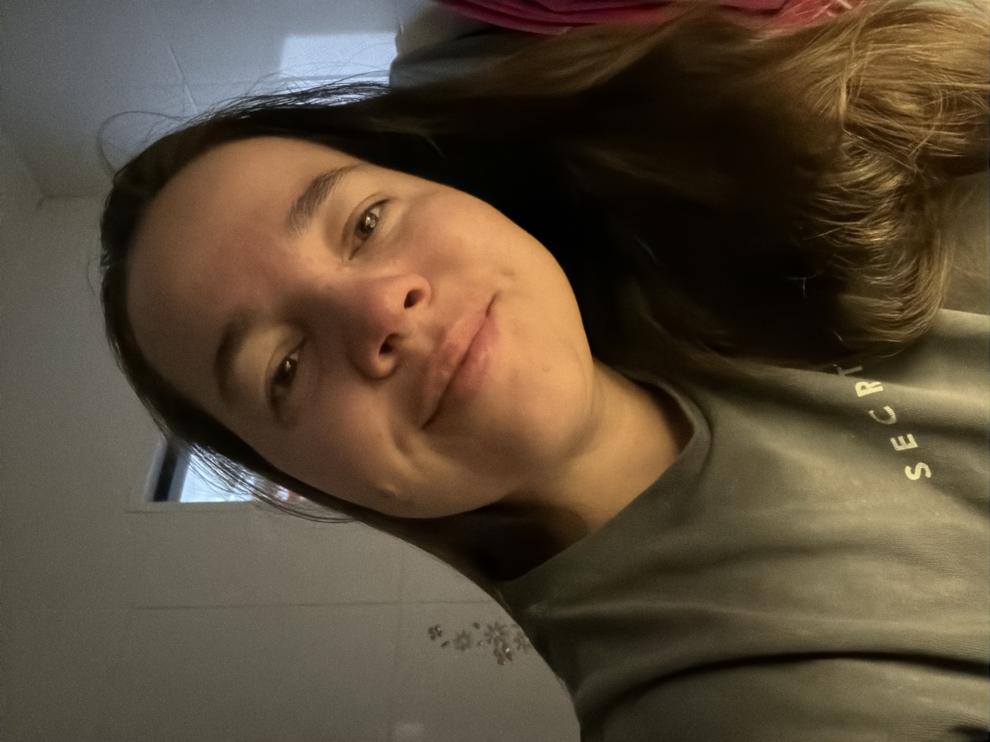 A selfie of a young woman with brown hair, fair skin, and no visible makeup. She is wearing a gray sweatshirt with the word "SECRET" partially visible on the upper right. The background is blurry and suggests an indoor setting, possibly a bathroom, given the glimpses of a tiled wall and a window with diffused light. The photograph provides no information to determine whether the location is in the city of Chaikovsky, Russia.