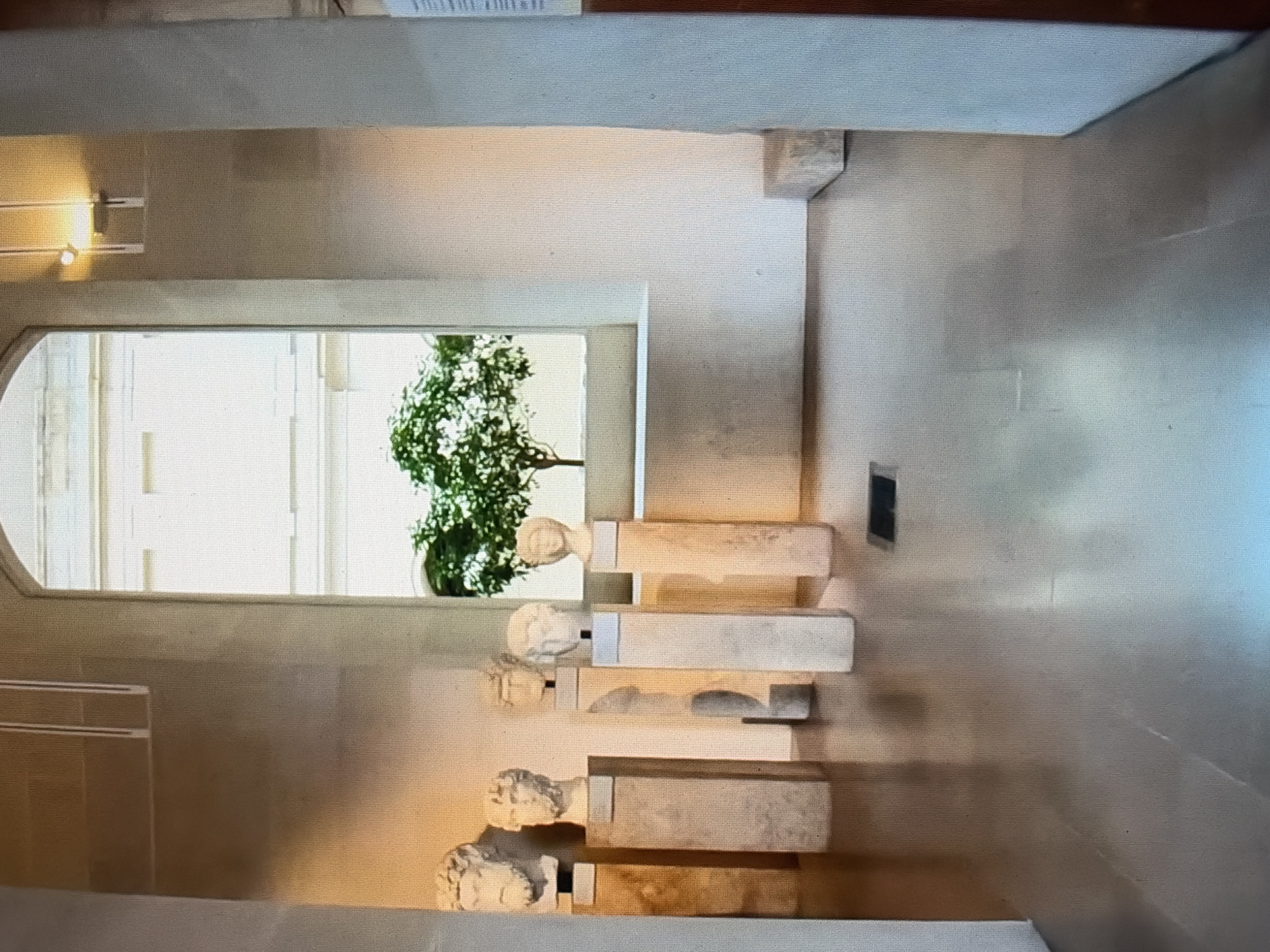 The image shows an interior view of a museum or gallery. In the foreground, the polished floor reflects the ambient lighting and the surrounding architecture. Several marble busts, each mounted on a tall, light-colored pedestal, are displayed. These busts are of various sizes and depict what appear to be ancient figures. In the middle ground, a large, arched window frames an outdoor scene featuring a lush green tree against a light-colored building. The wall to the right of the window is a neutral tone, and a small, dark rectangular vent is visible on the floor. The overall impression is one of quiet contemplation and artistic display within a structured architectural space. The location is unknown.