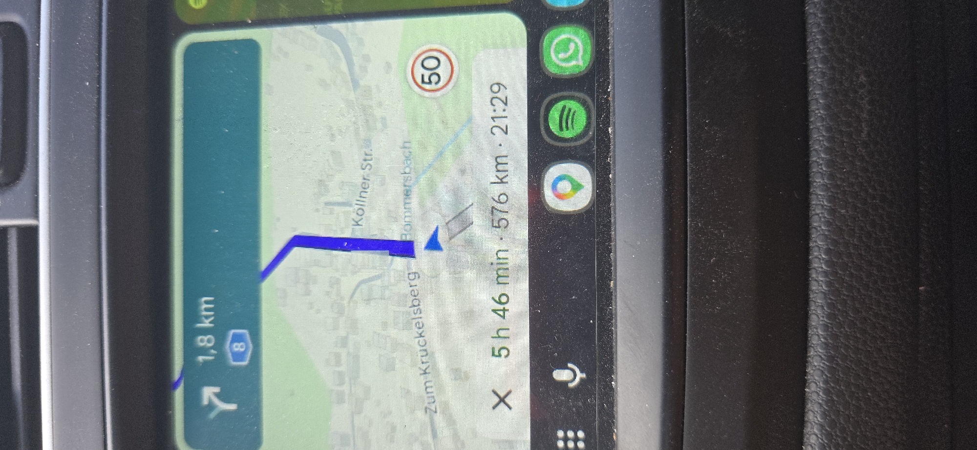 The image displays a navigation screen, likely from a car's infotainment system, showing a map with a route highlighted in blue. The screen indicates a distance of 1.8 km to the next turn, with the number 8 in a blue shield, suggesting a highway or main road. The route appears to be heading towards "Zum Kruckelsberg." Below the map, a journey summary shows an estimated time of 5 hours and 46 minutes and a total distance of 576 km, with an arrival time of 21:29. A speed limit sign for 50 km/h is visible on the right side of the map. At the bottom of the screen, icons for Google Maps, Spotify, and WhatsApp are displayed, along with a grid icon and a microphone icon, indicating potential app shortcuts. The screen is mounted in a car dashboard, with the texture of the dashboard visible. The context of Schwalbach, Germany is provided as the location.