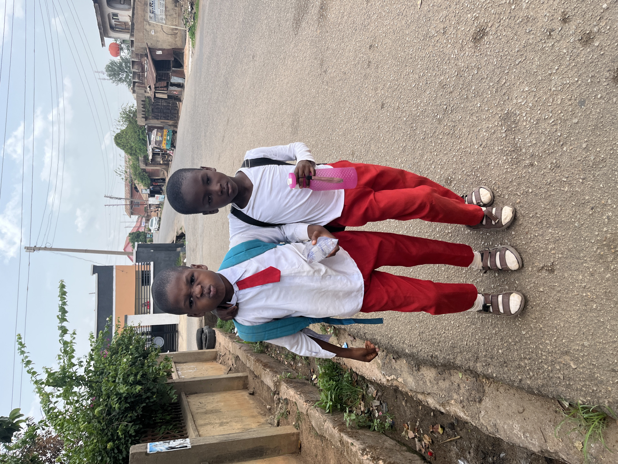 Two young boys in school uniforms stand on a paved road next to a curb in Jos, Nigeria. They are wearing white long-sleeved shirts, red trousers, and sandals with white socks. One boy has a blue backpack and a red tie, while the other carries a pink water bottle. The street is lined with buildings and vegetation. The sky is overcast.