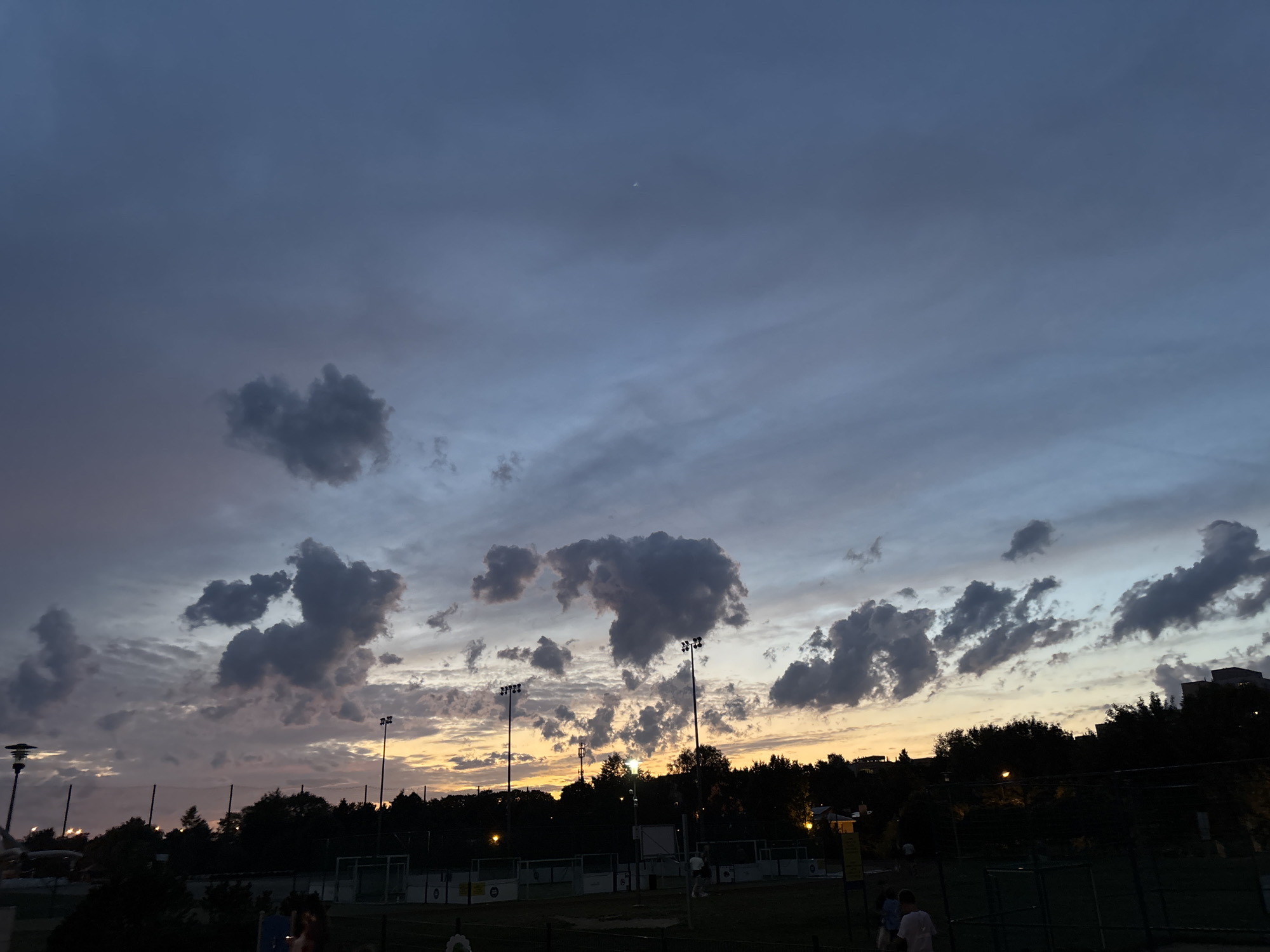 This is a wide-angle, dusk photograph taken from an elevated viewpoint, looking towards a sports field and a treeline at the city of Lublin, Poland. The sky is filled with dramatic, dark grey clouds, tinged with pink and orange from the setting sun near the horizon. A faint crescent moon is visible in the upper portion of the sky.
In the foreground, a dark silhouette of a fence runs horizontally across the bottom of the image. Beyond the fence, a sports field with artificial turf is visible, possibly a futsal or small soccer pitch, with white markings and low fencing. Several tall floodlights are positioned around the field, casting warm yellow light downwards. In the background, a dense line of dark trees forms a silhouette against the colorful sky. Behind the trees, buildings are partially visible, some with illuminated windows.
A few scattered individuals are visible on and near the sports field. Two people, one wearing a white shirt, are standing in the lower left corner of the frame. Another person in a white shirt is visible on the sports field towards the center right. The scene suggests a recreational activity occurring as evening approaches. The overall atmosphere is peaceful and serene, dominated by the striking twilight sky.