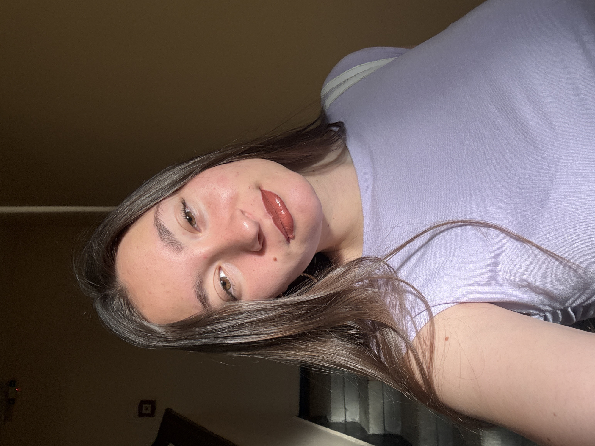 A young woman with long, dark brown hair is taking a selfie in a dimly lit indoor space. She is wearing a light purple top and her face is partially illuminated by sunlight coming from the left. A staircase is visible in the background to the left, and the walls are a warm, tan color. There is a small, dark square object on the wall in the upper left quadrant of the image. There is no visible indication of her location in Sofia, Bulgaria.