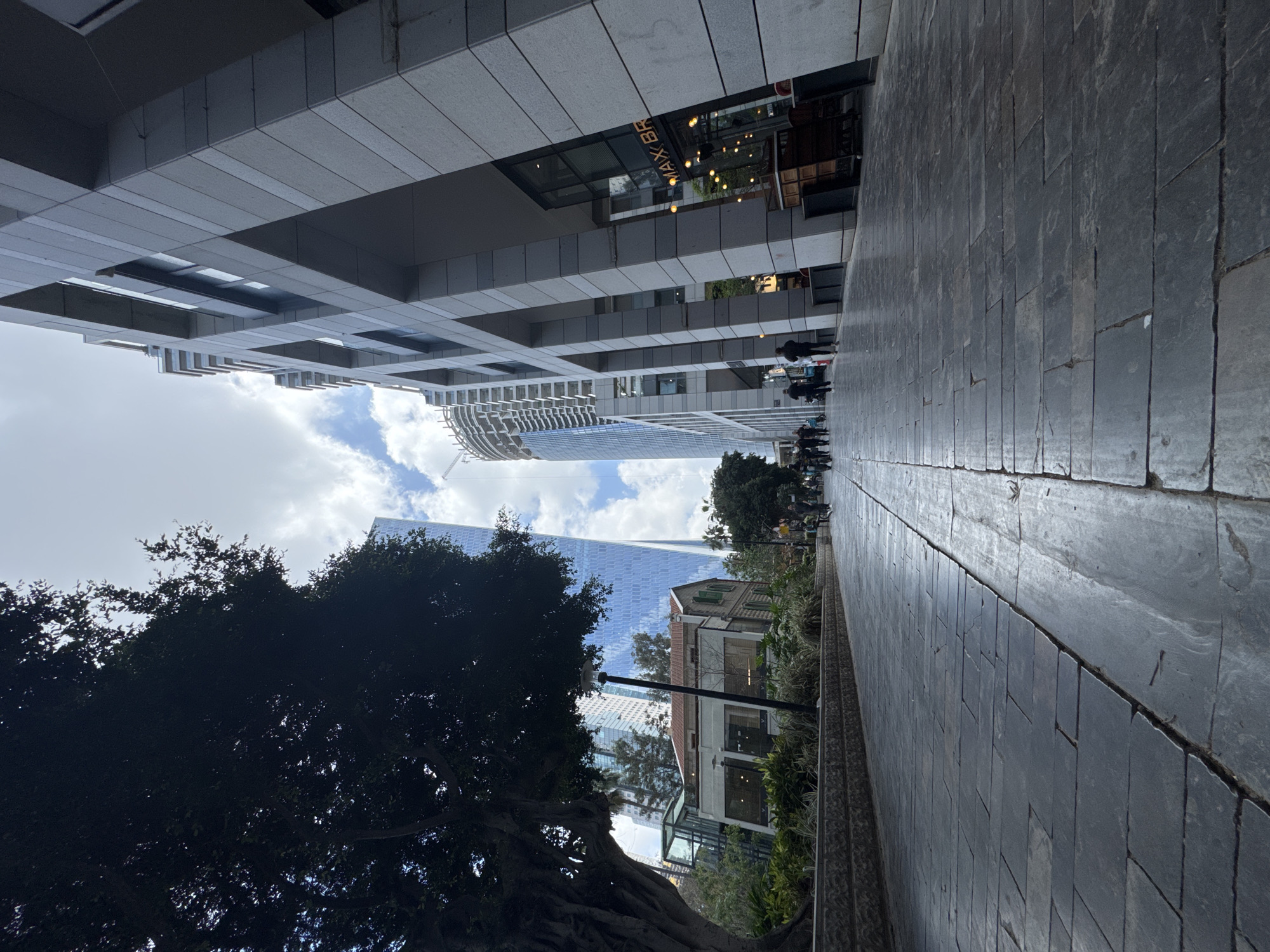 This image captures a vibrant urban street scene in Tel Aviv, Israel, showcasing a blend of modern and traditional architecture under a partly cloudy sky. The perspective is from ground level, looking down a wide paved pathway.
On the left side of the frame, several modern buildings rise. One prominent structure features a contemporary, multi-story design with horizontal concrete panels and glass sections. Further back, two sleek skyscrapers dominate the skyline; one has a distinctive curved, ribbed facade, while the other is a tall, reflective glass tower. A commercial sign reading "MAX BR" (likely "MAX BRENNER") with illuminated letters is visible on the first modern building.
The right side of the image features a dark grey paved walkway made of rectangular stone slabs, where several casually dressed people are seen walking. Lush green vegetation, including bushes and a large, dark-leafed tree with visible roots, borders the pathway. The tree partially obscures an older, quaint building with a red-tiled roof and green shutters, offering a sharp contrast to the towering modern structures. A black street lamp post stands near this older building.
The sky is partly cloudy with patches of blue visible, suggesting daytime. The artificial lights of the "MAX BR" sign are on, possibly indicating late afternoon or a dull day. The scene depicts a typical day in a bustling city environment, with pedestrians moving along the path.