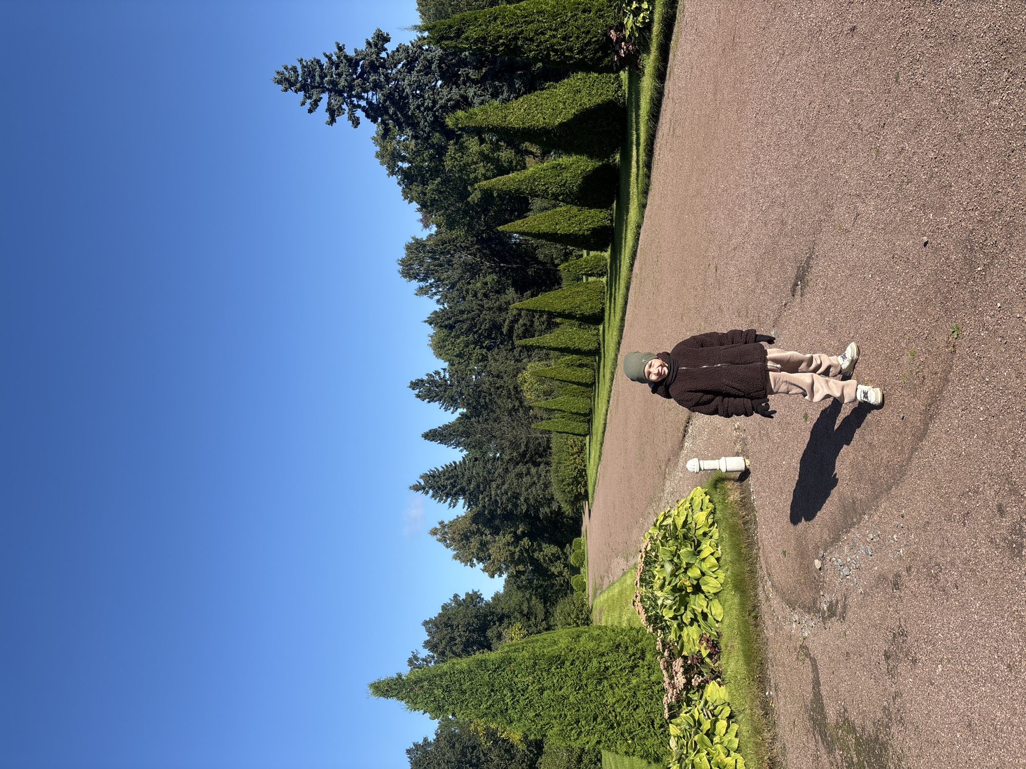 The image captures a clear, sunny day in the city of Peterhof, Russia. A young child, wearing a green beanie, a brown fleece jacket, beige pants, and white sneakers, stands on a gravel path in what appears to be a park or garden. The child is smiling and looking towards the camera. Behind the child, a well-maintained gravel path curves through a landscape of meticulously manicured green trees and bushes. To the left of the path, a bed of green plants with pink flowers is visible. The background features a dense line of various evergreen trees, including tall, cone-shaped cypresses and a large, mature pine tree on the right. The sky above is a vivid, cloudless blue. The scene suggests a peaceful, outdoor setting, likely within a public park or estate in Peterhof, known for its grand palaces and gardens.