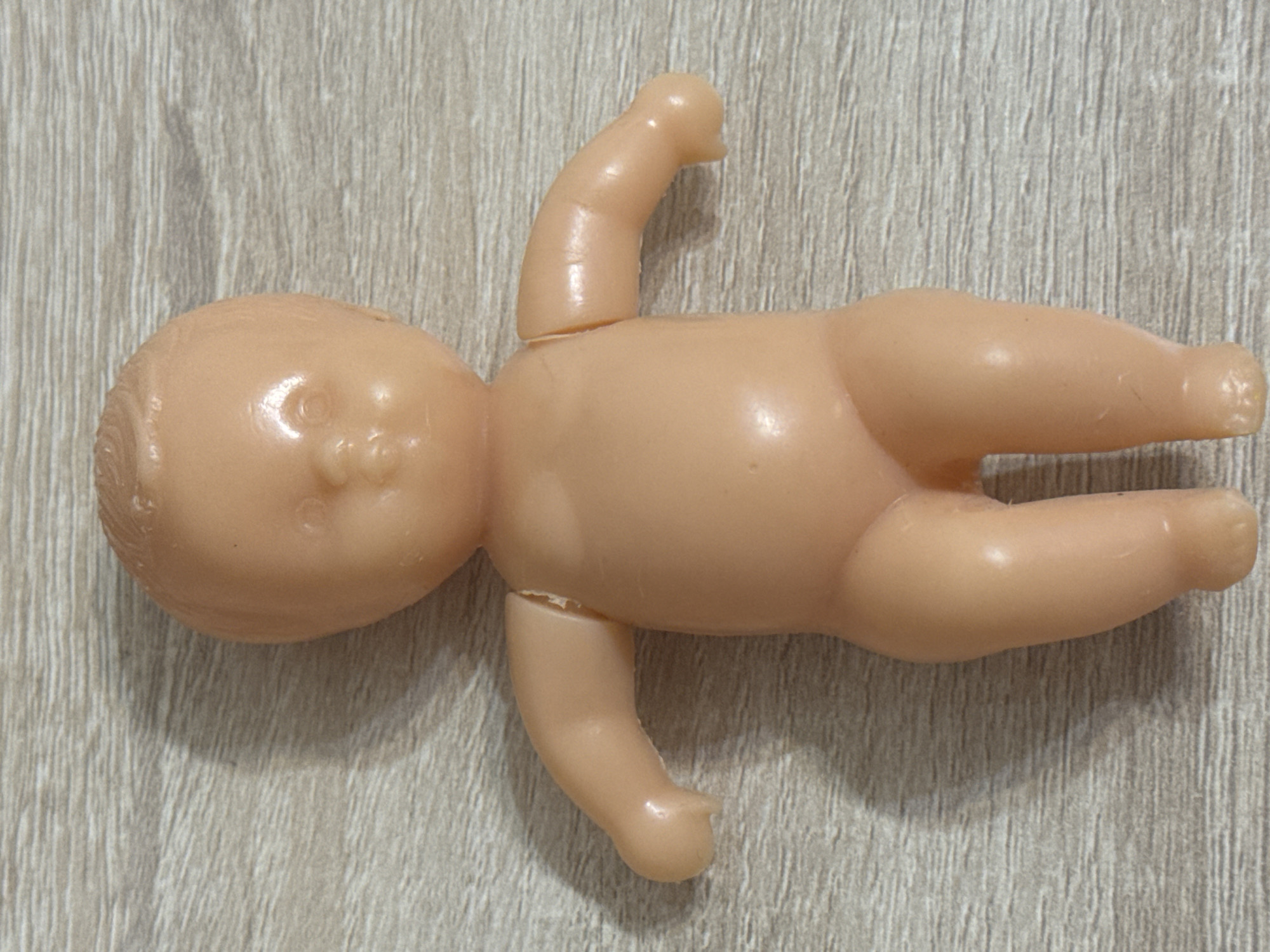 A nude, peach-colored plastic doll lies on its back on a light brown, horizontally-grained wooden surface. The doll has a rounded head with sculpted facial features, including closed eyes, a small nose, and slightly parted lips. Its body is plump with a defined torso, arms, and legs. The arms are outstretched to the sides, and the legs are bent at the knees. Both arms appear to be detached at the shoulder joint, with visible gaps where they connect to the torso. The doll is positioned in the center of the frame, with a shadow cast to its left. The background is consistent throughout the image, with no discernible urban or environmental features.