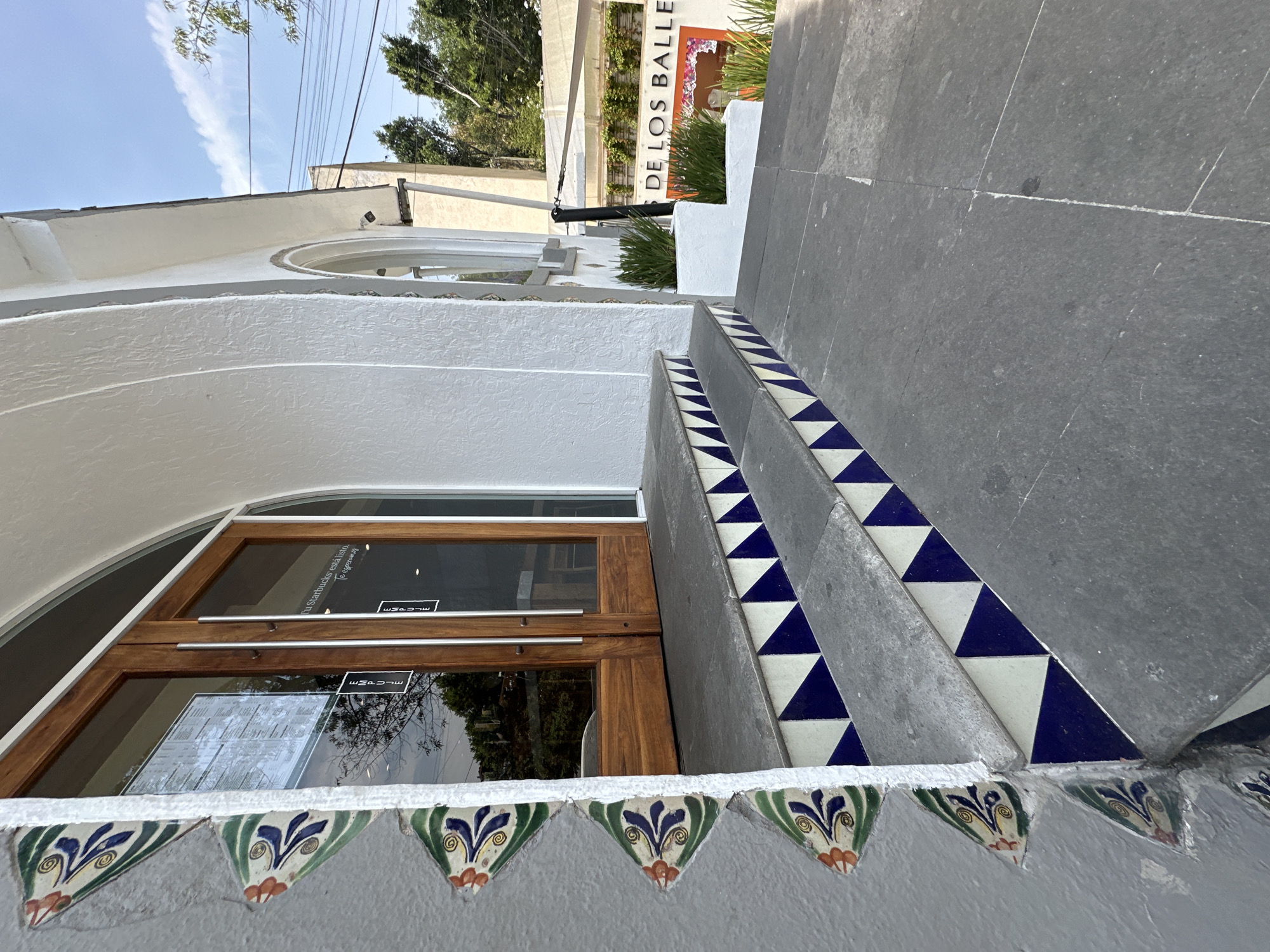 This image presents a low-angle view of a building entrance in Mexico City, bathed in bright sunlight on a clear day. The foreground features a set of outdoor steps crafted from dark gray stone. The risers of these steps are distinctively decorated with alternating blue and white triangular tiles, creating a bold, geometric pattern. Below the steps, a white stucco wall is adorned with a border of colorful ceramic tiles, each spade-shaped with intricate green, blue, and orange floral or foliate designs.
A warm-toned wooden double door with large glass panels serves as the entrance. Reflections on the glass reveal surrounding trees and the sky. Text visible on the glass reads, "Tu Starbucks está listo. Te esperamos" (Your Starbucks is ready. We'll wait for you), indicating the presence of a Starbucks outlet or a location selling its products. A smaller, reversed text, possibly "ESCAPADE," is also faintly visible on the glass.
In the upper background, part of a sign displays the text "DE LOS BALLE." Overhead utility wires crisscross the bright blue sky. Lush green trees and foliage are visible beyond the building, adding natural elements to the urban scene. A small security camera is mounted on the building's white facade in the upper right. The architectural style, characterized by white stucco and vibrant decorative tiles, reflects a distinctive regional aesthetic. No individuals are present, lending a quiet, inviting atmosphere to the entrance.