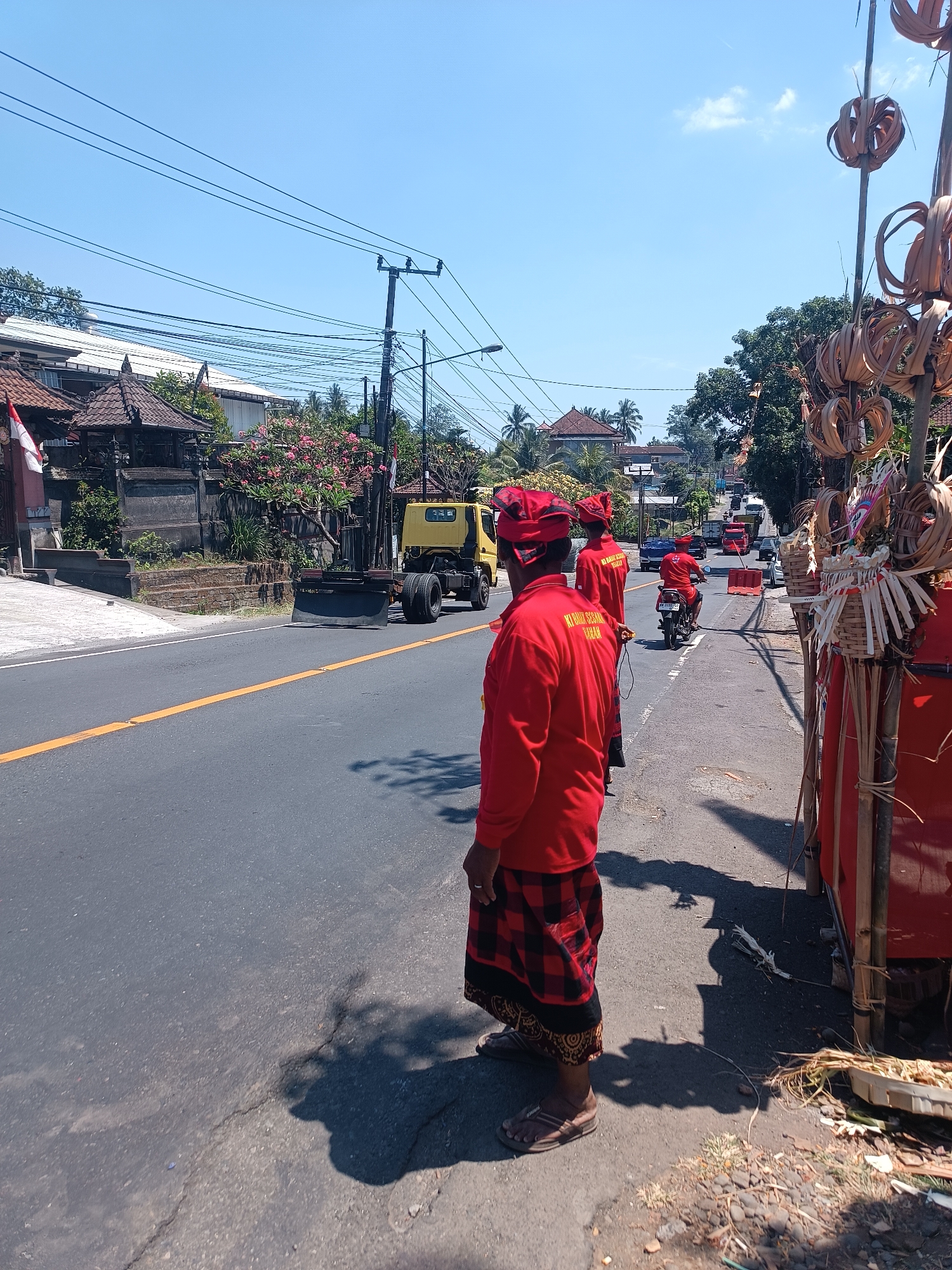 The image depicts a street scene in Kerambitan, Indonesia, under a clear blue sky with minimal cloud cover. In the foreground, three individuals dressed in traditional red attire with red head coverings are standing on the side of a paved road. The person closest to the camera is facing away, showing the back of their red shirt with text on it, and a black and red checkered sarong. The other two individuals are also facing away or partially away from the camera, also wearing red shirts and sarongs.
The road has a yellow line marking the center. On the left side of the road, there are buildings with tiled roofs and some vegetation, including trees with pink flowers. A yellow truck with a large blade attachment on the front is parked on the side of the road. Further down the road, traffic can be seen, including cars and motorcycles, and traffic cones are placed on the road.
On the right side of the image, there are tall bamboo poles adorned with woven, circular dried palm leaf decorations. A red structure, possibly a stall or a vehicle, is partially visible behind these decorations. The ground on the right is unpaved with scattered dry grass and debris. The overall scene suggests an event or gathering taking place on a public road, with some form of traffic management or decoration present.