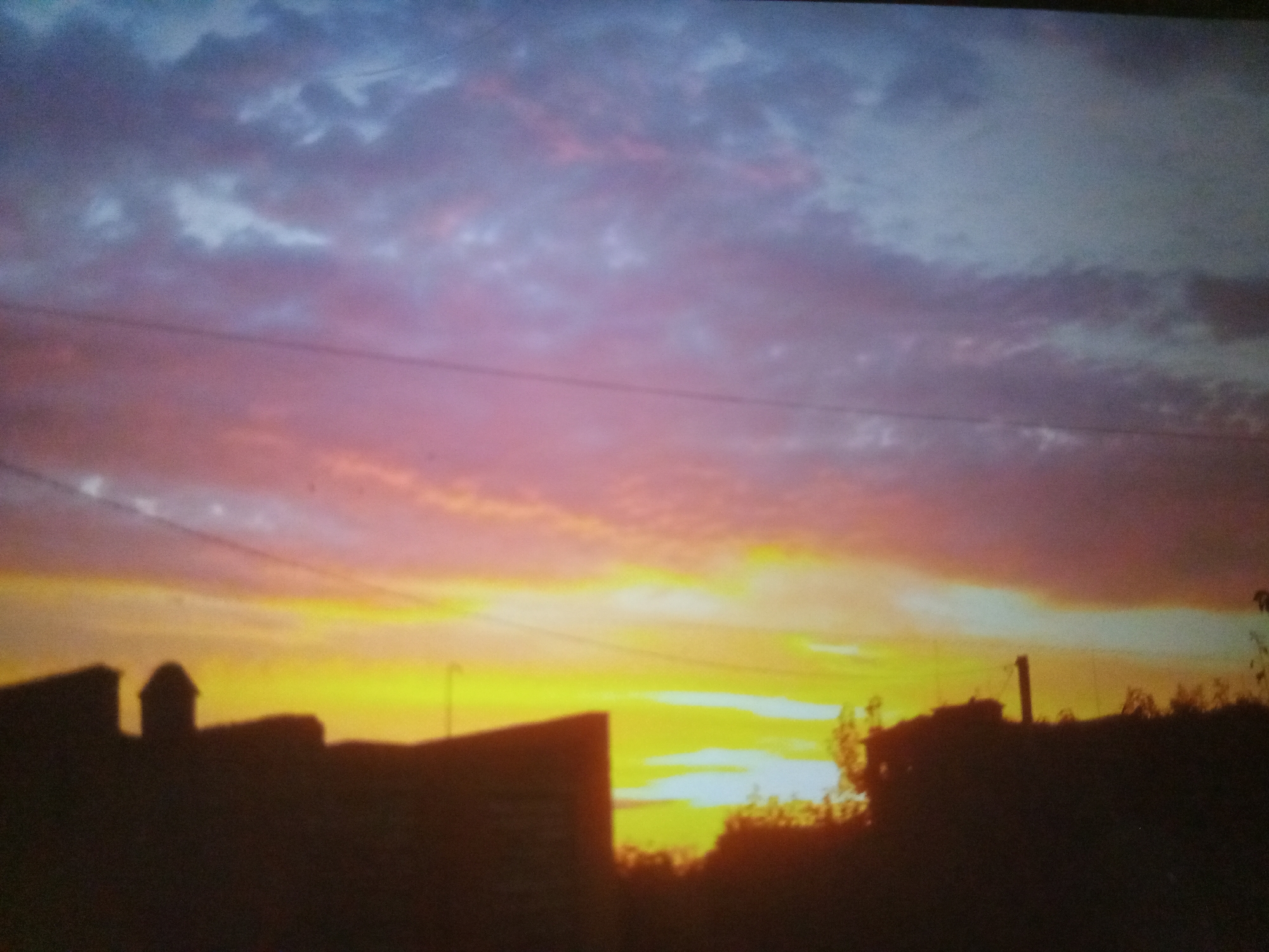 This is a photograph of a sunset over the city of Myrhorod, Ukraine. The sky is filled with dramatic clouds in shades of pink, purple, and yellow, indicating the sun is low on the horizon. In the foreground, the silhouettes of buildings and trees are visible against the vibrant sky. One prominent silhouette appears to be a domed structure, possibly a church or part of a larger building complex. Another silhouette to the right suggests utility poles or similar structures. The overall scene is one of a serene and colorful end to the day in an urban or suburban setting.