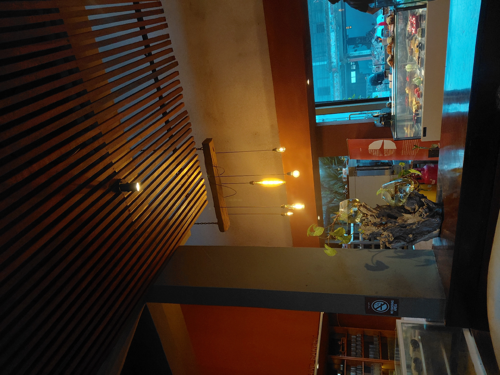 The image depicts the interior of a cafe with a modern and rustic design. The ceiling is adorned with parallel wooden slats, and multiple Edison-style light bulbs hang from a wooden beam. A column stands prominently in the foreground, bearing a "No Smoking Area" sign. To the left, shelves are filled with glassware, and a display case showcases various pastries. To the right, a large window offers a view of the street, with a display counter filled with more desserts. A person is partially visible near the window, possibly a staff member. The overall ambiance is dimly lit with warm, inviting tones. The location is identified as a cafe in Unknown, Unknown.