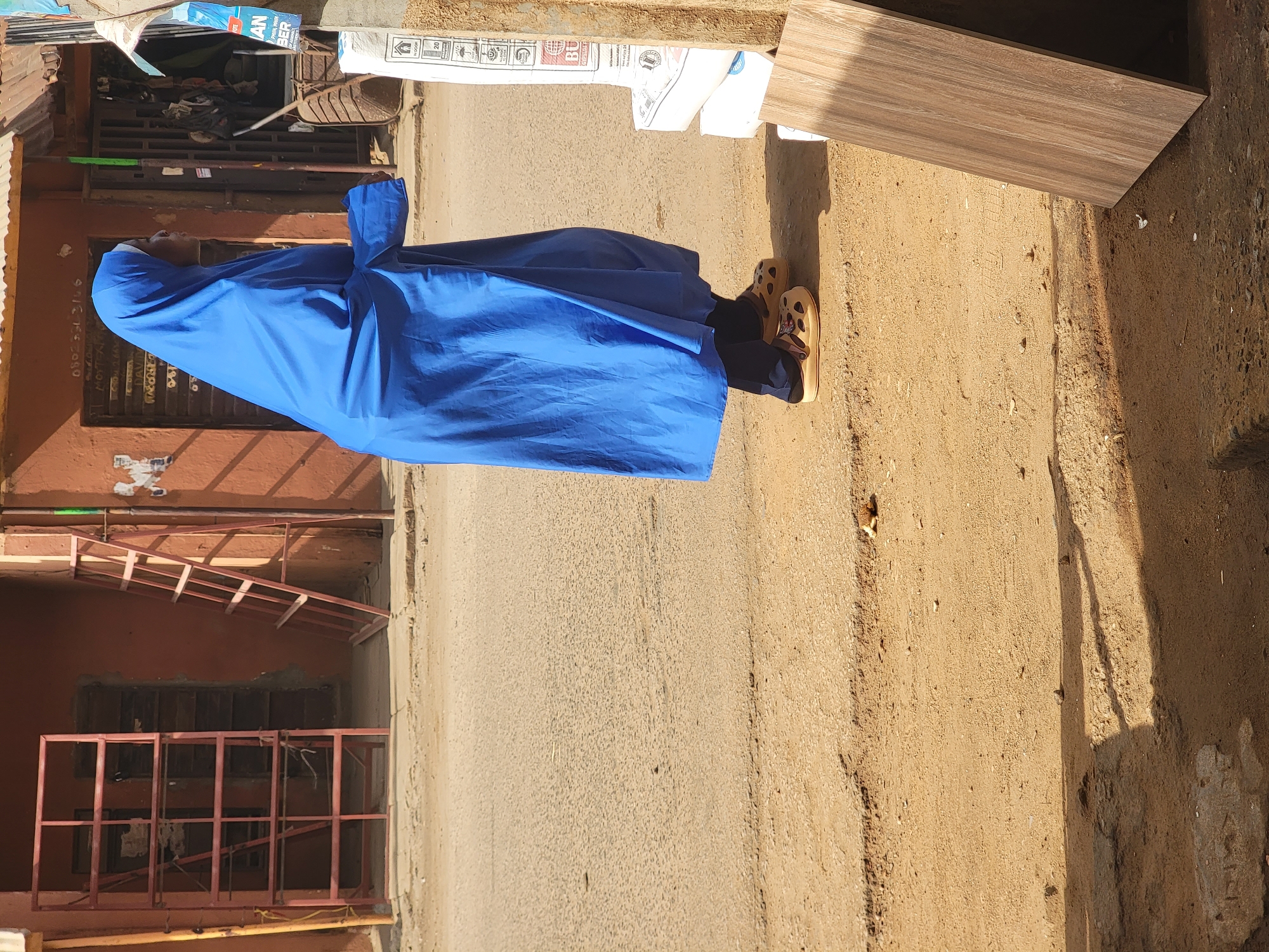 An individual clad in a blue garment stands on a paved surface alongside a street in Maiburiji, Nigeria. The surrounding environment includes structures with metal security features and a partially visible wooden panel or object.