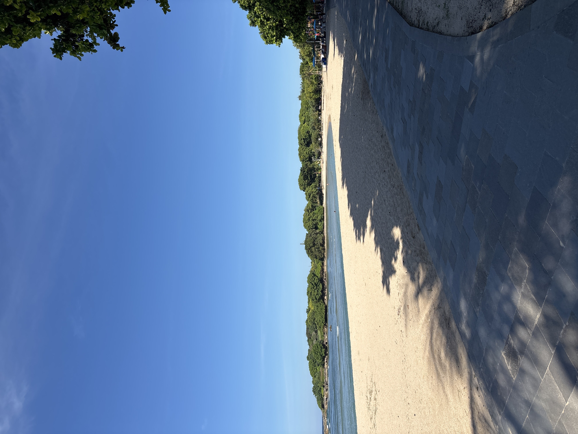 The image captures a beach scene in Unknown, Indonesia, on a clear day. The sky is a vibrant blue, mostly unobstructed by clouds. A line of lush, green trees borders the beach. The sand is light in color, and there are shadows cast by the trees. Next to the beach there is a tiled pathway. The water, presumably the sea, runs parallel to the beach, with some visible activity of people enjoying the water.