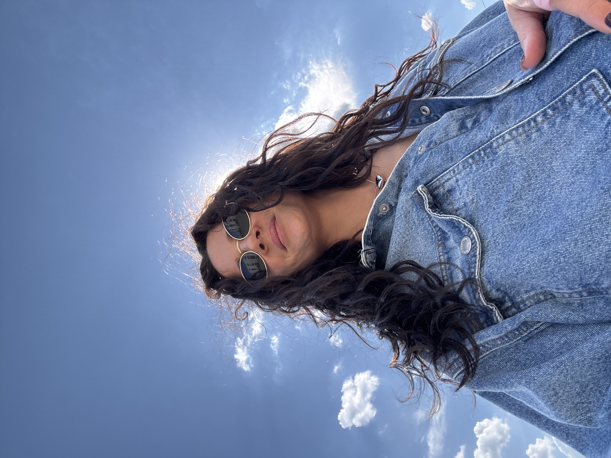 A woman with long, dark, curly hair is wearing sunglasses and a denim jacket. The sun is behind her head, creating a flare. The sky is blue with scattered clouds. The city of Chaikovsky, Russia, cannot be determined from the image.