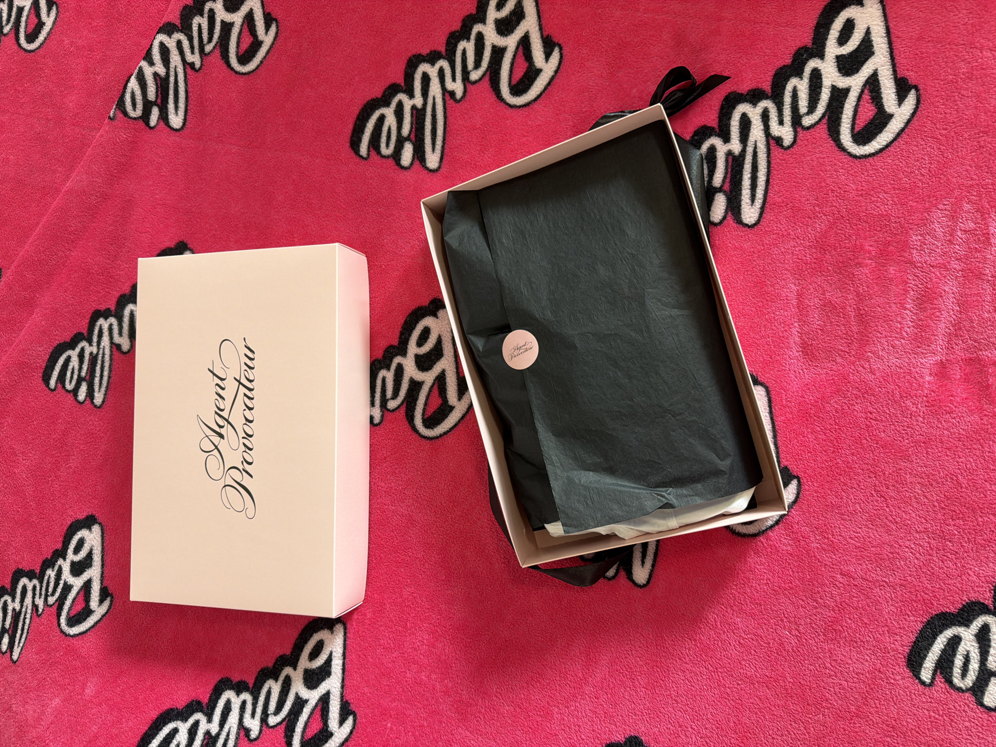 The image depicts two boxes on a pink Barbie-themed blanket. One box, labeled "Agent Provocateur," is closed. The other is open, revealing black tissue paper and a black ribbon. There is no indication of the location being Moscow, Russia.