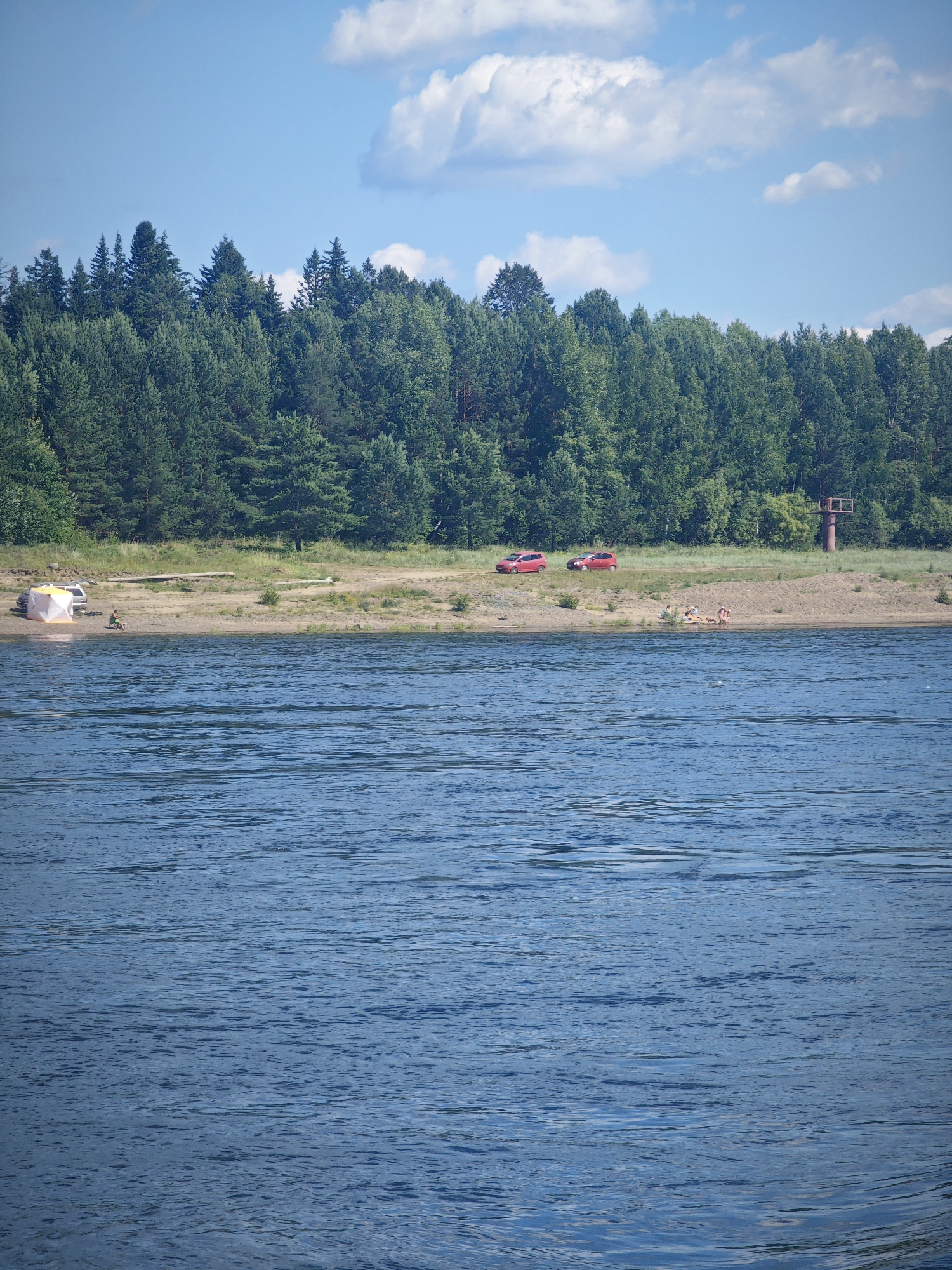 The image depicts a sunny day at a body of water, likely a river or lake, in the vicinity of Abalakovo, Russia. In the foreground, the water's surface is rippled, reflecting the blue sky and scattered white clouds.
Along the shore in the middle ground, a sandy bank is visible, backed by a dense forest of evergreen trees. On the sandy bank, several objects and individuals are present. Two red cars are parked on the grass verge near the trees. Closer to the water, a pop-up tent with white and orange panels is set up, with a single person sitting nearby. Further along the beach, a small group of people are gathered on blankets, some appearing to be sunbathing or relaxing. To the far right of the sandy area, a red metal tower structure stands. The overall environment suggests a recreational area by the water.