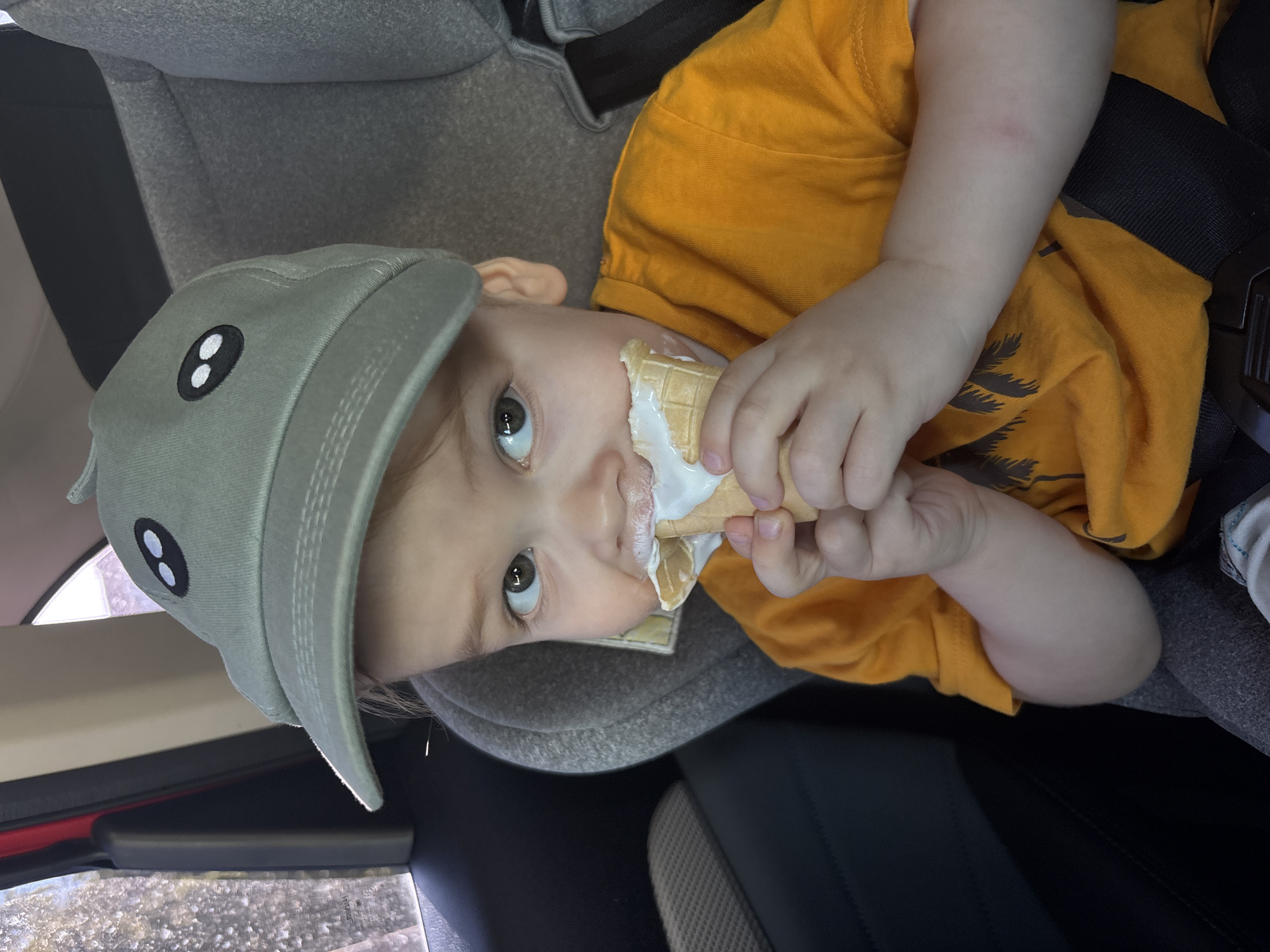 A young child is sitting in a car seat, wearing a grey cap with two embroidered white-outlined black ovals, and an orange short-sleeved t-shirt with dark brown palm tree graphics. The child is holding an ice cream cone with melting white ice cream smeared around the mouth. The car seat has grey upholstery and a black safety harness. The interior of the car is visible, with dark grey and black elements. Based on the available information, the location cannot be determined.