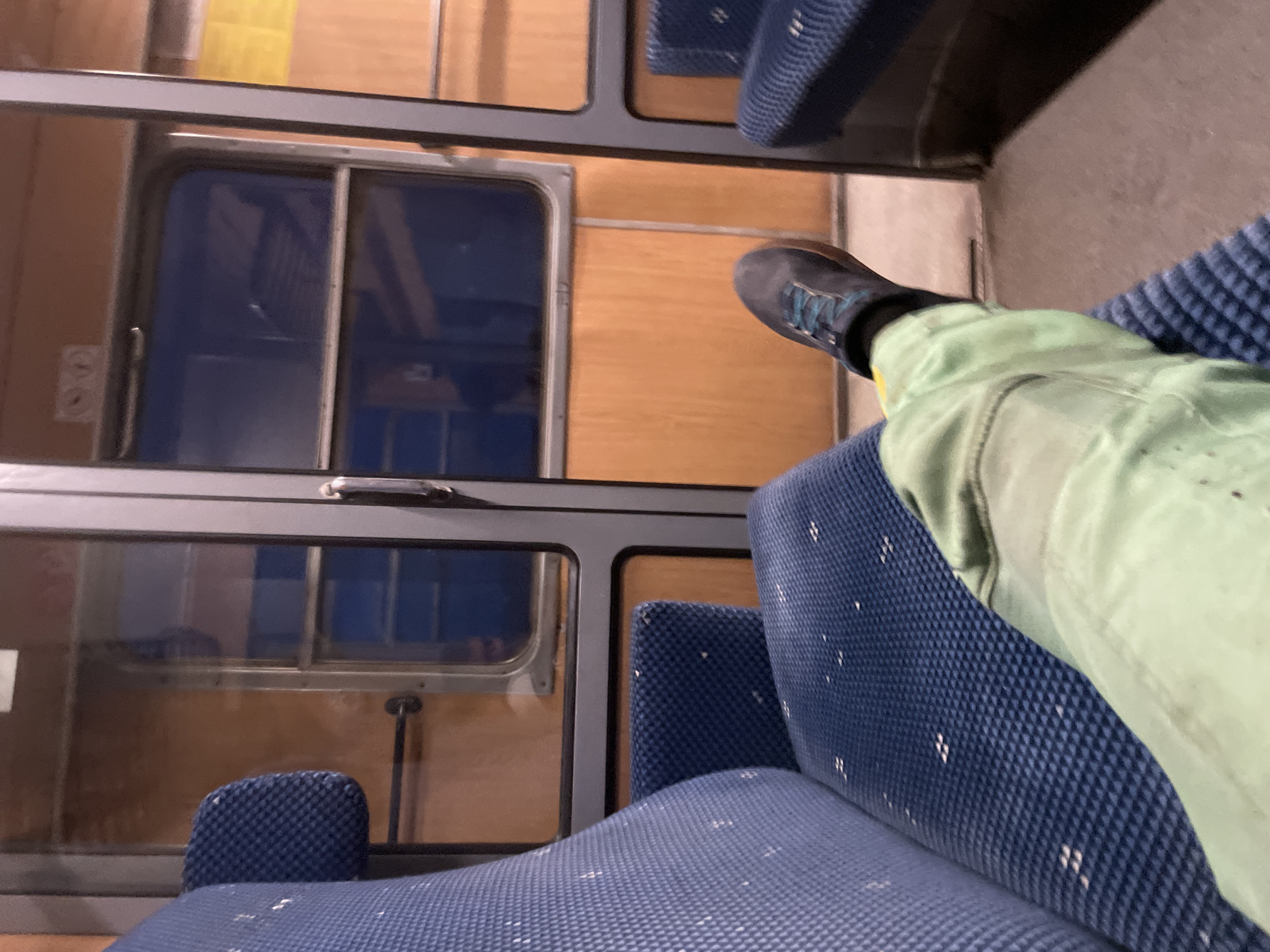 The image shows the interior of a train carriage, with a focus on a person's legs and feet sitting on a blue upholstered seat. The train compartment features wooden paneling on the walls and large windows that reflect the interior. The person is wearing light green trousers and dark blue shoes with blue laces. The environment suggests a mode of transportation, likely a train, given the seating arrangement and the windows. The crops provide close-up views of the seat upholstery, the train doors, and the person's footwear, aiding in detailed visual analysis. The image does not provide any explicit information or context related to the city of Borovo, Bulgaria.