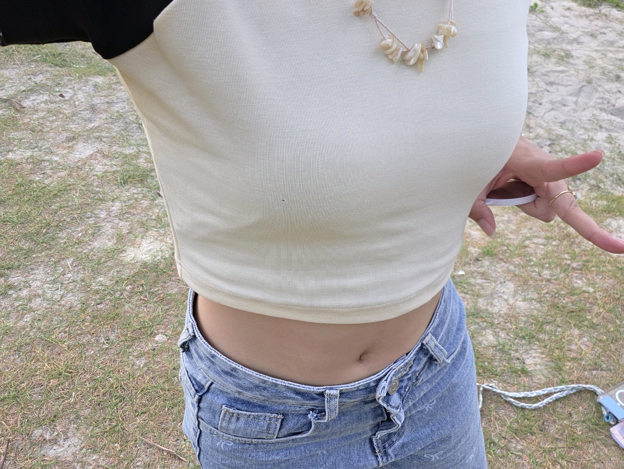 A person wearing a cream-colored crop top and blue denim jeans stands outdoors. The crop top has black sleeves and a necklace made of shells around the neck. The person's midriff is exposed, and they are wearing a gold ring on their left ring finger. Their right hand is reaching towards a white object, possibly a phone, with their index finger extended. The background consists of dry grass and sandy ground. The scene is set in George Town, Malaysia.