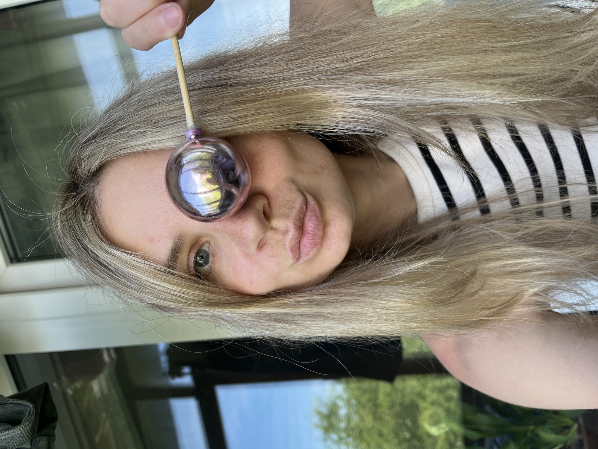 A blonde woman with light skin is holding a reflective purple ornament attached to a wooden stick in front of her left eye. She has a neutral expression and is wearing a striped shirt. The background shows a window with a reflection of trees and sky, and a glimpse of a building exterior suggesting an outdoor setting. The photo appears to be taken in Chaikovsky, Russia.