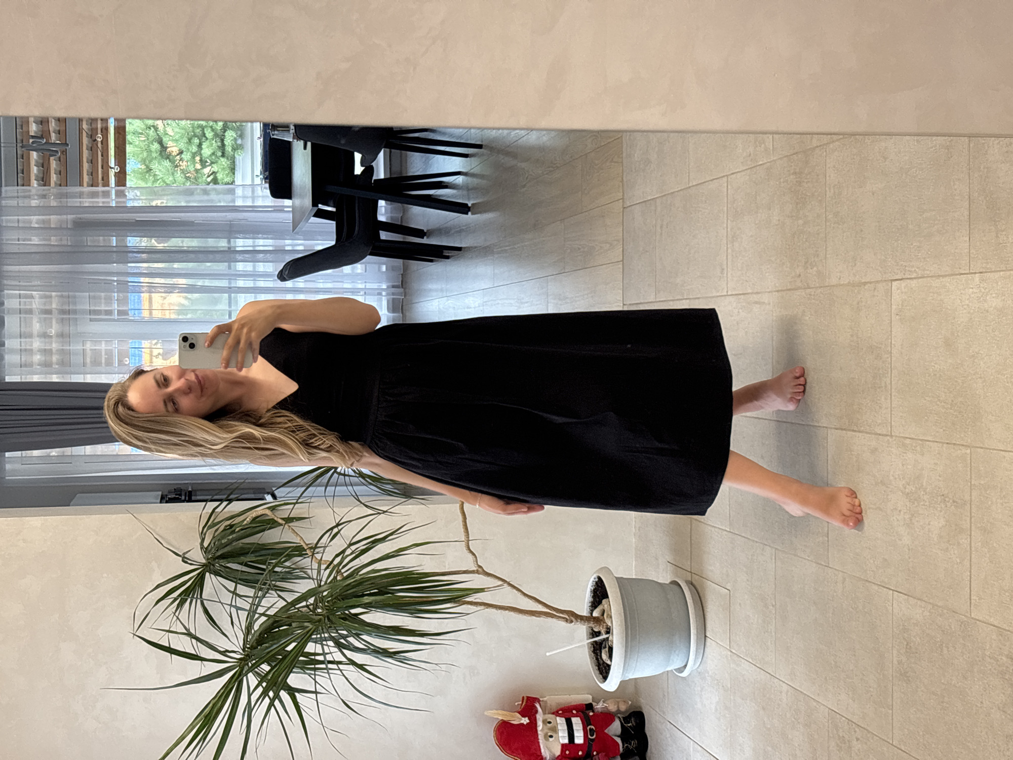 A woman with blonde, wavy hair, wearing a black V-neck dress and barefoot, stands in an indoor setting, taking a mirror selfie. To her left, a tall potted plant with long, spiky leaves is situated near a decorative nutcracker dressed in a red and gold uniform. The floor is tiled with large, beige squares. In the reflection of a mirror to the right, a dining area is visible with a dark table and chairs, and sheer curtains covering a window that looks out onto greenery. The wall behind the plant has a textured finish. The overall environment appears to be a modern, well-lit domestic interior, possibly in Chaikovsky, Russia, given the context.
