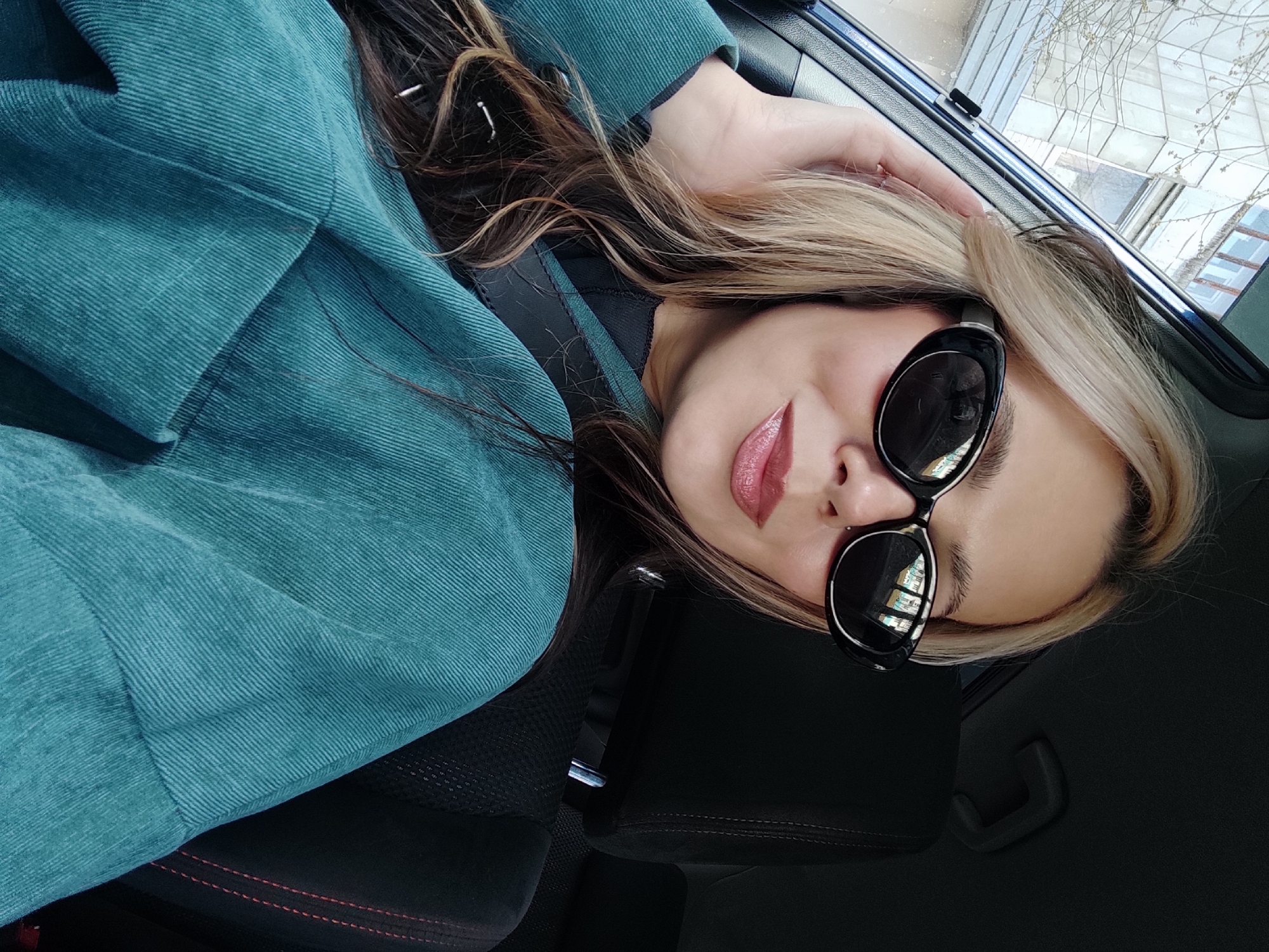 A woman is captured in a portrait-style image, appearing to recline or lie back in a car seat. The image is rotated, placing her head towards the top right. She has long, wavy hair with blonde highlights mixed with darker strands, flowing around her head and shoulders. Her right hand rests gently on her hair near the car window.
She is wearing large, black-framed sunglasses that cover her eyes. The lenses reflect indistinct patterns of what appears to be buildings and a bright sky. Her lips are visible, painted with glossy, pinkish-red lipstick, and slightly parted. She is dressed in a teal or turquoise corduroy jacket or coat, characterized by its prominent ribbed texture. A black car seatbelt is visible across her chest.
The interior of the car is primarily black, with subtle red stitching visible on the side of the seat. Through the car window, bare tree branches are visible against a bright, possibly overcast, sky, suggesting a late winter or early spring setting. Faint outlines of distant structures can also be discerned. The scene depicts a casual, daytime moment inside a vehicle, located in Chaikovsky, Russia. No visible text is present in the image.