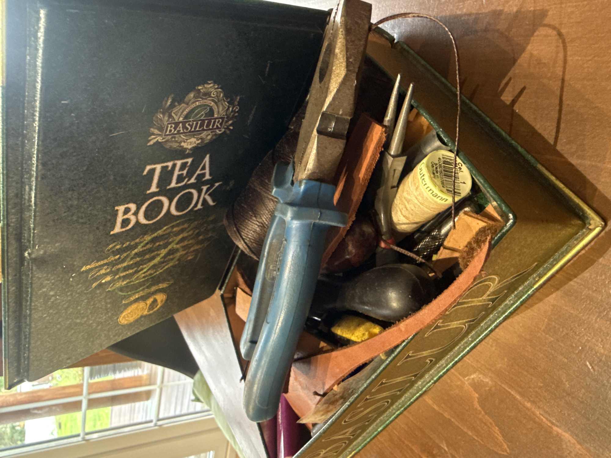 The image shows a close-up view of an open, antique-style tin, possibly a tea tin, filled with various crafting tools and materials. The tin's exterior is dark green with embossed gold lettering that reads "TEA BOOK BASILUR". Inside, there are pliers with blue handles, spools of thread (one with a "Gutermann" label), leather scraps, a pointed awl-like tool, and other small metal implements. The tin is placed on a wooden surface, likely a table. The background shows a blurred view of windows and greenery, suggesting an indoor setting near a natural outdoor environment. There are no people or animals present in the image. The scene depicts a collection of tools for crafting or repair, organized within a decorative tin.