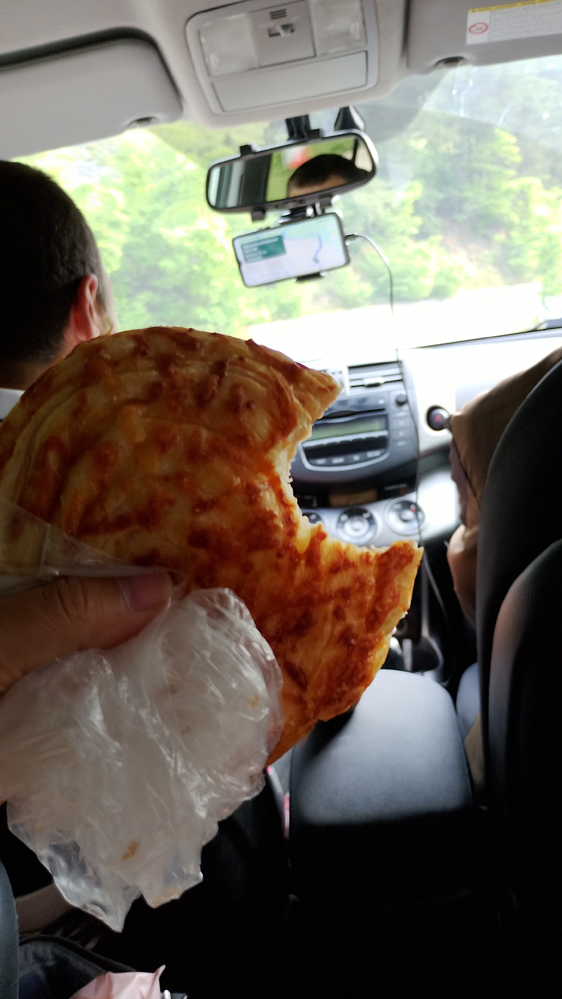 The image, taken inside a vehicle, features a person's hand holding a piece of pizza wrapped in plastic. The pizza appears to have a cheese topping and a bite taken out of it. The background shows the interior of a car, including the dashboard, a rearview mirror with a phone mounted on it displaying a navigation app, and the heads of the driver and another passenger. Outside the car, greenery is visible, suggesting a rural or natural setting. While the image itself does not provide direct evidence of the location being Hvoina, Bulgaria, the context suggests it was taken during a journey.