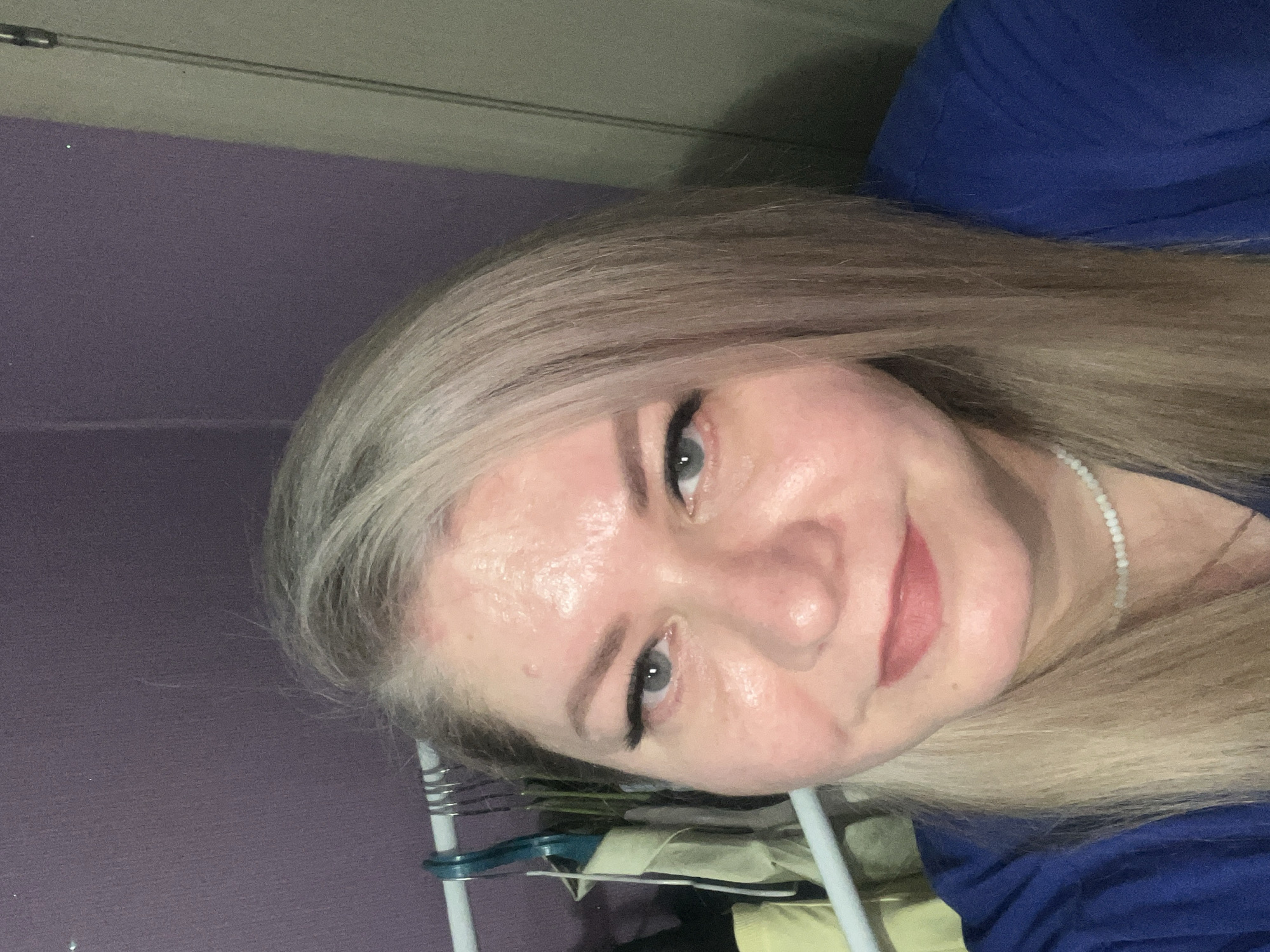 A woman with blonde hair and light skin is pictured in a close-up, rotated portrait. She is wearing a blue shirt and a beaded necklace. Her makeup includes eyeliner. Behind her is a pale purple wall and a clothes rack with garments hanging on it. There is no indication that the image was taken in Izhevsk, Russia.