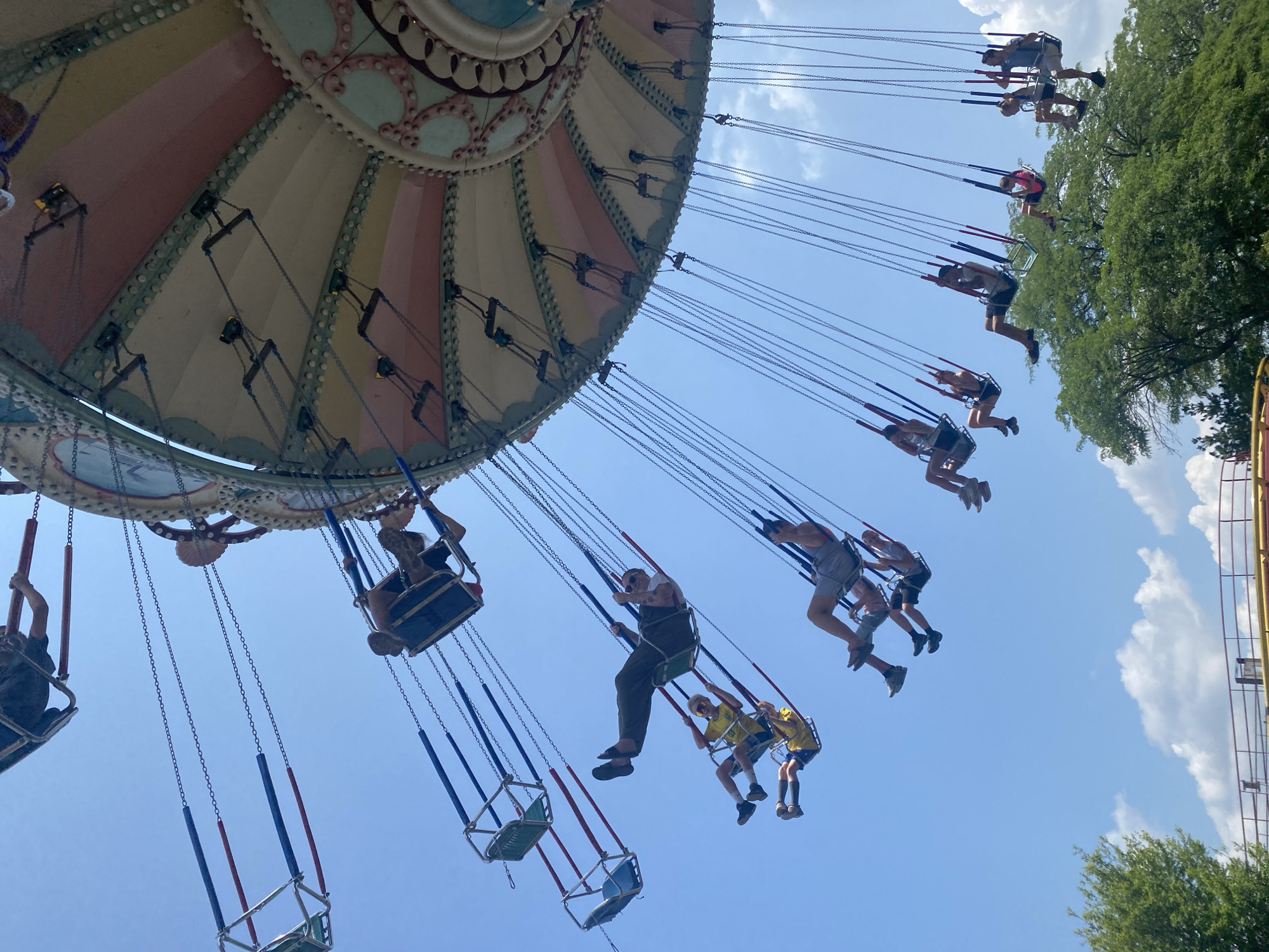 This image captures a thrilling ride on a swing carousel, likely at an amusement park, under a bright blue sky dotted with scattered white clouds. The carousel's decorative top, adorned with pastel colors and intricate patterns, is visible at the top right, with numerous chains descending from it, each suspending a seat. Several individuals are seated on these swings, some appearing to be adults and others possibly teenagers, actively enjoying the ride as they are propelled outward and upward. The subjects are dressed in casual summer attire. In the lower portion of the frame, lush green trees and the upper railing of a structure are visible, suggesting the ride is situated within a park environment. Without further context or external information, it is not possible to definitively pinpoint the location to Montreal, Canada.
