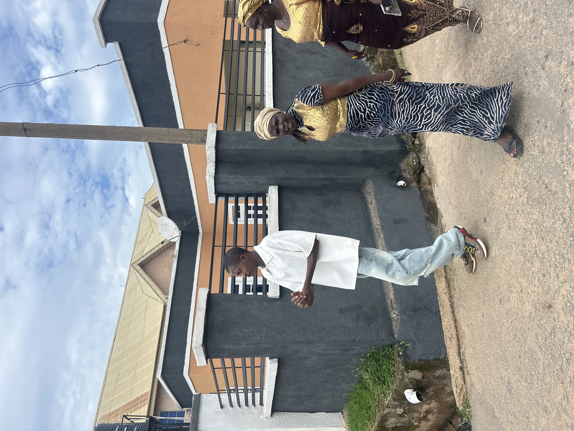 The image displays a street scene in Jos, Nigeria, featuring three individuals walking along a paved path adjacent to a building complex under a partly cloudy sky. On the left, a male individual of apparent young adult to middle age, with a slight mustache, wears an oversized white collared short-sleeved shirt, light blue distressed jeans with a tear on the left knee, and brown, black, and yellow sneakers. He walks with his hands clasped together, looking slightly to his left. To his right and slightly behind him, a middle-aged woman with dark complexion wears a light beige headwrap, a gold sequined short-sleeved top, and a long skirt with a black, white, and silver abstract pattern. She has gold bangles on her right wrist and wears black flat sandals, looking towards the camera. To her right, a second middle-aged woman, also with dark complexion, wears a gold headwrap, a gold sequined top, and a dark brown long skirt with intricate gold embroidery. She wears white low-heeled sandals, a yellow ring on her right ring finger, and holds a dark rectangular object in her left hand, looking forward and slightly left. All three individuals are walking in the same general direction, from right to left across the frame.
The walking surface is a textured, light gray asphalt or concrete. To the immediate left of the path, a low dark gray concrete wall with a white coping runs parallel to the path, featuring black horizontal metal railings. Behind this wall, a building with sections of orange-tan and white facade is visible. A black water tank on a stand is partially visible at the far left, behind the wall. A white satellite dish is mounted on the orange section of the building's facade. Above the building, a large corrugated metal roof, light gold or tan in color, extends across the upper background. A concrete utility pole rises vertically on the right side of the frame, with dark, thin wires hanging from it. Near the base of the wall on the far left, a patch of green grass grows, and a concrete drainage ditch with a white pipe opening is present.