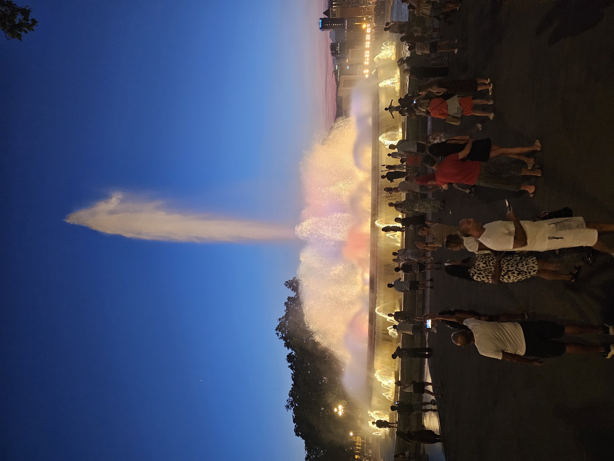The image captures a lively evening scene at a fountain show in Barcelona, Spain. A large crowd of people are gathered to watch the illuminated water display, which features vibrant colors and impressive jets of water shooting high into the twilight sky. The sky transitions from a deep blue at the top to softer hues of pink and orange closer to the horizon, indicating it is dusk or early evening.
The fountain itself is the central focus, with water illuminated by colorful lights, creating a magical atmosphere. Many people are holding up their phones to capture the spectacle, suggesting excitement and engagement with the event. The crowd is diverse, with individuals and groups of various ages. The setting appears to be an outdoor plaza or park, with trees visible on the left side of the frame and buildings in the background, illuminated by the fountain's glow and city lights. The overall mood is festive and awe-inspiring, as people come together to enjoy the visual performance. The ground is dark, suggesting it is a paved area.