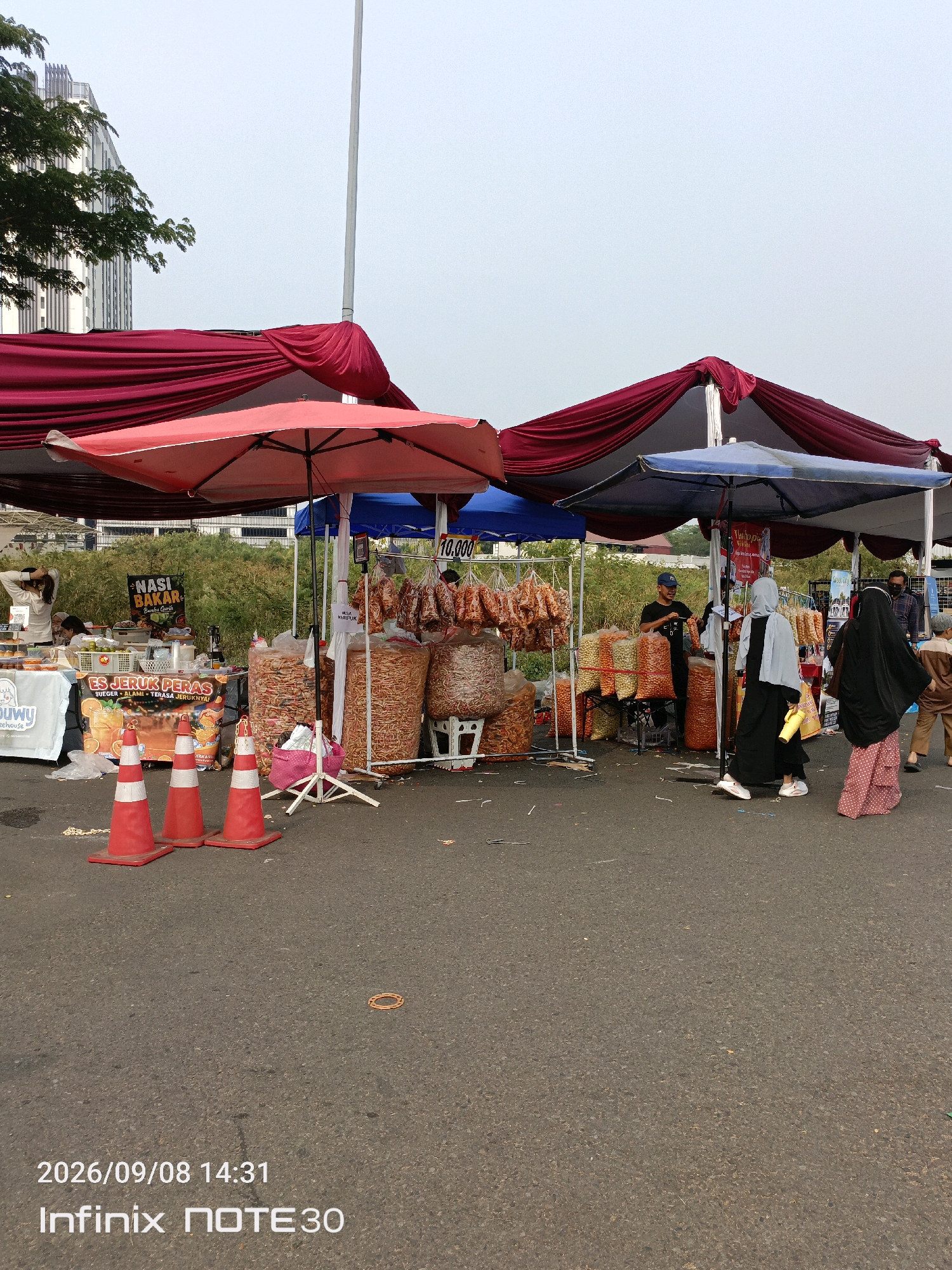 The image depicts an outdoor market scene in East Jakarta, Indonesia. Several stalls are set up under large red and blue awnings, with a backdrop of trees and a modern building. The foreground shows a paved area with traffic cones, suggesting it might be a temporary market set up on a road or open space.
The stalls are selling various food items, primarily snacks and prepared foods. One stall features large bags of puffed snacks and packaged goods, with some items hanging on display. Another stall advertises "Nasi Bakar" (grilled rice) and "Es Jeruk Peras" (freshly squeezed orange juice).
Several people are present in the image. A vendor wearing a dark shirt and a blue cap is seen interacting with customers. A woman in a grey hijab and a black abaya is walking past the stalls, carrying a yellow bag. Another woman in a black hijab and a pink polka-dot skirt is also visible. A child in brown clothing is walking away from the camera. Other individuals are seen in the background, potentially shoppers or vendors.
The overall atmosphere suggests a bustling marketplace. The presence of a modern building in the background indicates an urban environment, likely on the outskirts of a more developed area. The sky is overcast, suggesting it's either early morning or late afternoon. The date and time stamp at the bottom of the image indicate it was taken on September 8, 2026, at 14:31.