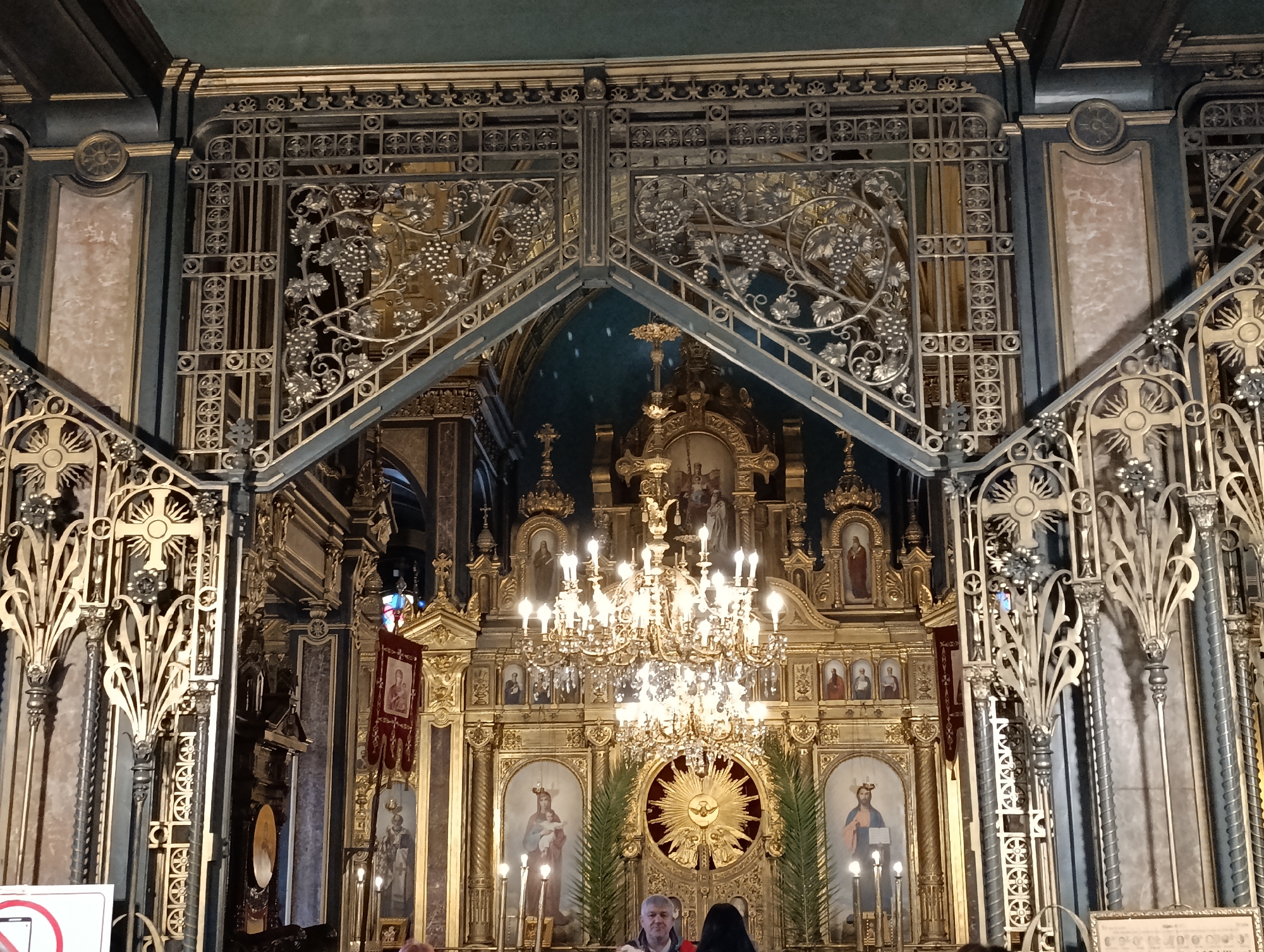 The image captures the interior of St. Stephen Bulgarian Orthodox Church (Iron Church) located in Istanbul, Turkey. The focal point is the ornate gold iconostasis, intricately decorated with religious figures and topped by a large chandelier. An elaborately patterned iron gate-like structure frames the view of the iconostasis. Two people are visible in the lower part of the picture.