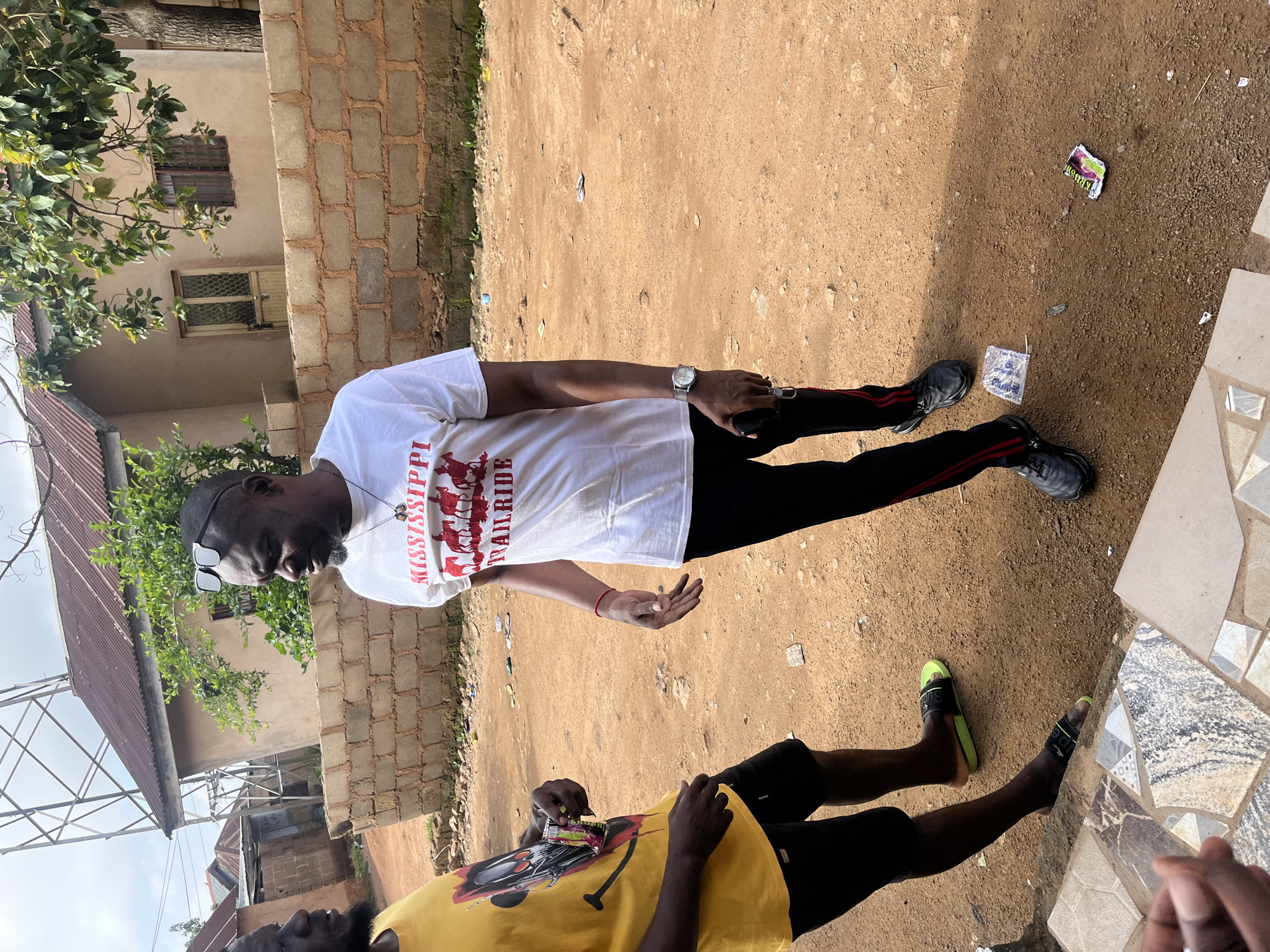Two men are standing outside in what appears to be Gura Top, Nigeria. One man, wearing a yellow t-shirt and black shorts, is holding a small, colorful packet. The other man, wearing a white t-shirt with red lettering that reads "MISSISSIPPI TRAILRIDE" and black pants with red stripes, is looking at the first man and gesturing with his hands. He has sunglasses perched on his head and is wearing a watch on his left wrist. The ground is dirt, with scattered debris. In the background, there are simple buildings with corrugated metal roofs and a brick wall. A tall metal structure, possibly a cell tower, is visible in the upper left corner. The sky is overcast.