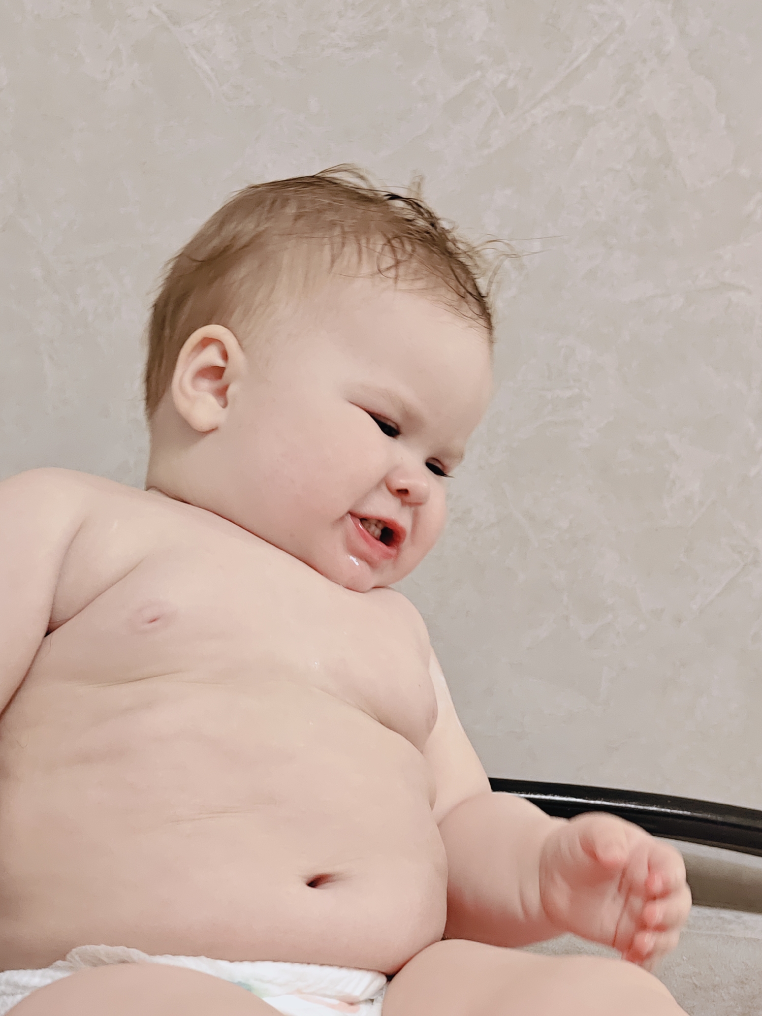 A close-up, low-angle shot captures a baby indoors, likely an infant or young toddler, in a setting that suggests a bath or being seated in a high chair. The baby has fair skin, light brown, slightly damp or messy hair, and is shirtless, revealing a plump torso with visible skin folds and a belly button. A diaper is visible at the bottom of the frame. The baby's expression is one of intense emotion, with eyes squinted, mouth open to reveal a few teeth, and a scrunched-up face, suggesting crying, fussiness, or strong reaction. Drool or water is visible around the baby's mouth and chin. The baby's right arm is slightly raised, with a closed fist. The background consists of a light beige or off-white wall with a subtle, textured pattern. A dark line, possibly the rim of a tub or chair, is present in the lower right corner. The lighting is soft and even, indicating an indoor setting in Chaikovsky, Russia. There is no visible text.