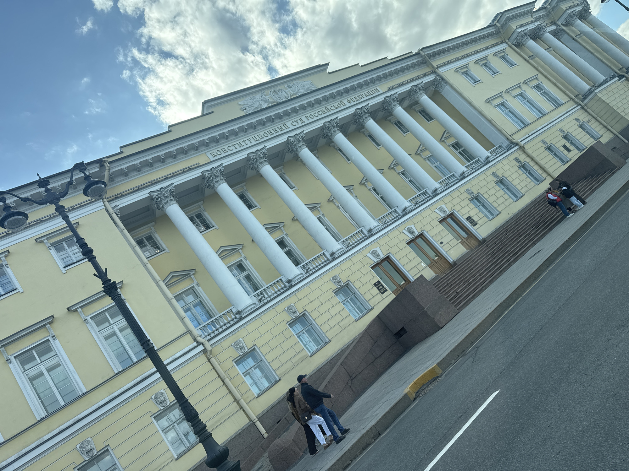 The image shows the Constitutional Court of the Russian Federation, located in Saint Petersburg, Russia. The building is a light yellow, neoclassical structure with prominent white Corinthian columns. An inscription is visible above the columns in Cyrillic: "Конституционный Суд Российской Федерации," which translates to "Constitutional Court of the Russian Federation."
Several people are present in the image. On the right, a small group of people are standing on the steps leading to the building's entrance. Another pair of people are walking along the sidewalk near the base of the building.
A black, ornate lamppost stands on the left side of the frame. The street is visible on the right, marked with a white line. The sky is blue with scattered white clouds.