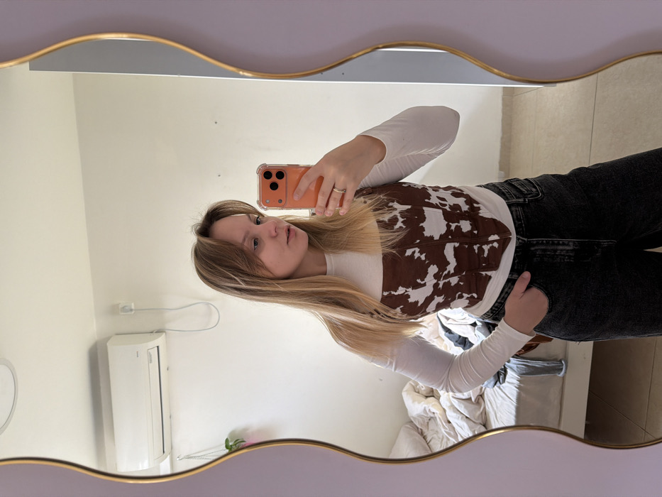 A young adult female is depicted taking a mirror selfie in what appears to be a residential bedroom. She has long, straight, light brown hair with a fringe and is looking slightly towards her left (viewer's right). Her attire consists of a white long-sleeved, high-neck top, a brown and white cow-print strapless corset-style top, and dark-wash, wide-leg jeans. A silver-toned ring is present on her left ring finger. She holds an orange-colored smartphone, encased in clear plastic, in her right hand, pointed towards the mirror to capture the image. Her left hand is placed inside the pocket of her jeans.
The mirror has a wavy, irregular light purple frame with a gold-colored inner edge. The reflection reveals a room with light-colored, possibly off-white, walls. A white wall-mounted split-system air conditioning unit is affixed to the wall in the upper left portion of the reflection. Below and to the left of the AC unit, a small potted plant with green foliage is suspended. To the lower left, an unmade bed with a white frame is visible, covered with white and light-colored bedding, along with some dark fabric items. Light-colored tile flooring is discernible at the bottom of the reflected image. An electrical outlet with a white cable is visible on the wall behind the subject's head. The environmental cues are consistent with a typical residential interior in Tel-Aviv, Israel.