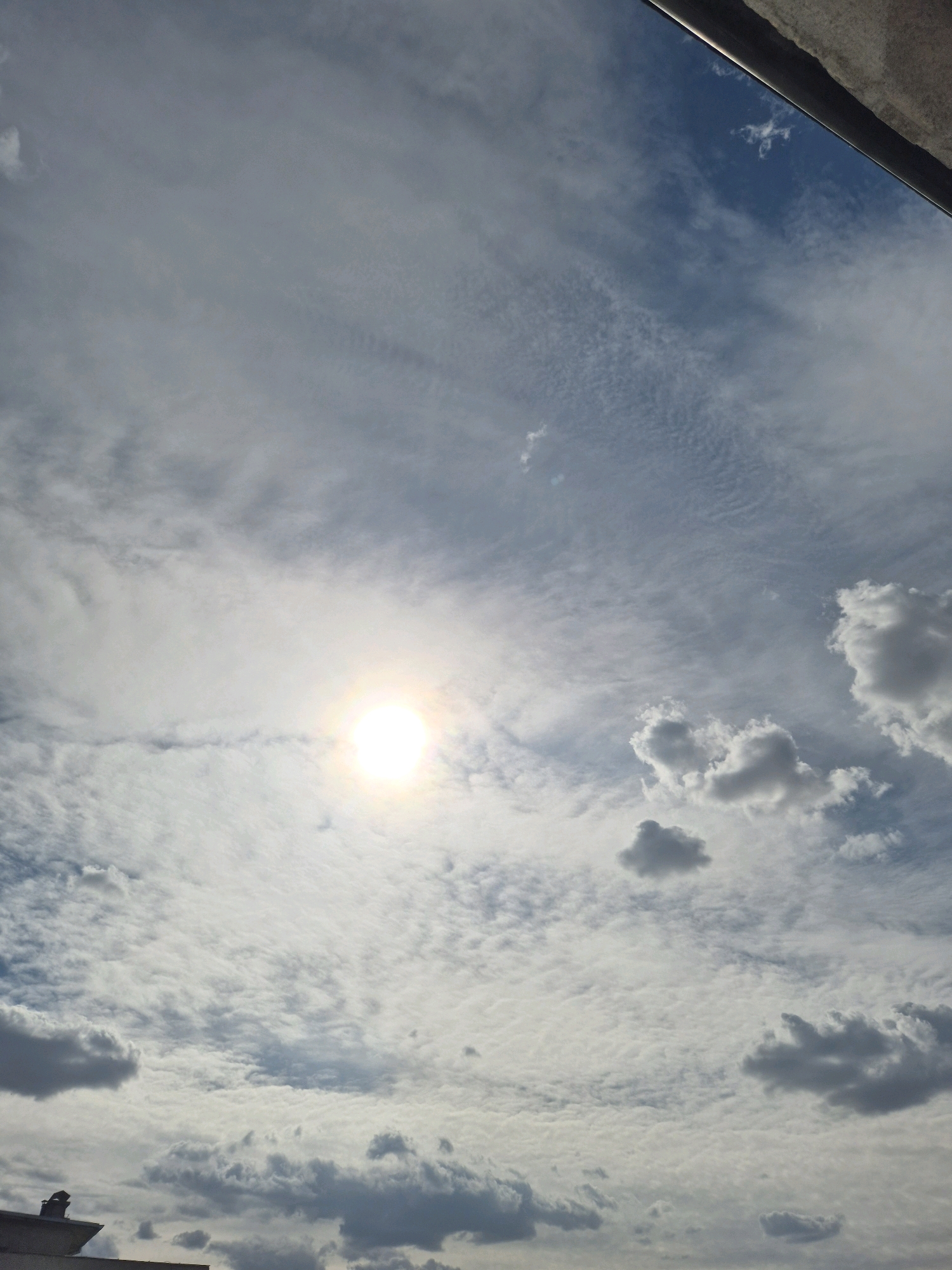 A bright sun is visible in the sky, partially obscured by thin clouds. The sky is blue and filled with scattered clouds of various sizes and densities. Some clouds are wispy and spread out, while others are more defined and puffy, with some appearing darker, suggesting they are thicker or catching less light. The overall impression is of a partly cloudy day. The image also captures a portion of a building's edge in the lower left corner, with a dark, angled structure resembling a roof or ventilation system. In the upper right corner, a metallic edge, possibly part of a building or structure, runs along the top of the frame.