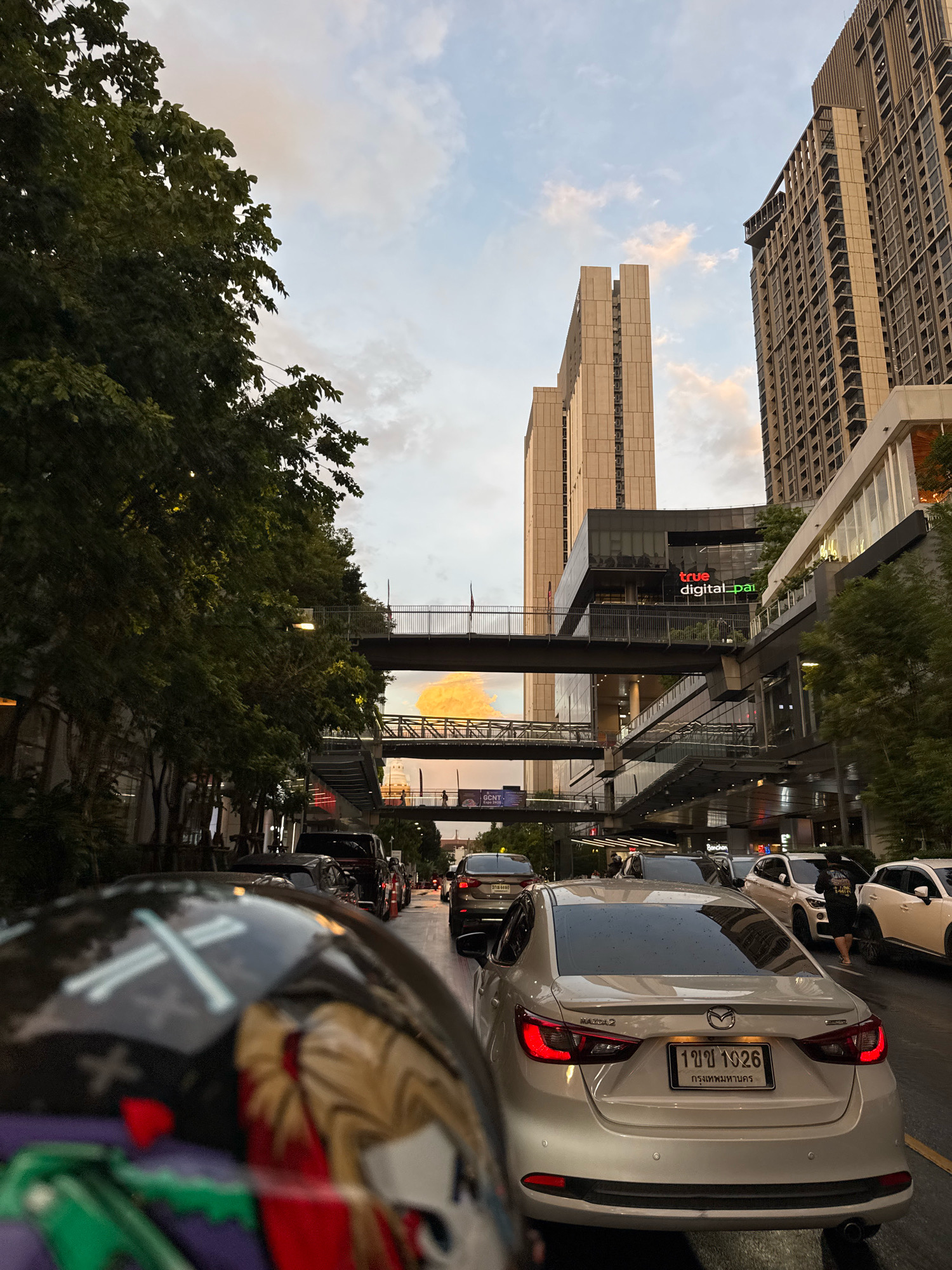 The image depicts a street scene in Bangkok, Thailand, likely during a traffic jam. The perspective is from the rider of a motorcycle, looking forward at the traffic ahead. In the foreground, the motorcycle's helmet is partially visible, featuring a design with white lines forming a stylized cross.
The main focus of the image is the heavy traffic on the road, with several cars lined up. The most prominent vehicle is a silver Mazda 2, clearly showing its license plate which reads "1 ขจ 1926" and "กรุงเทพมหานคร" (Bangkok). Other vehicles, including SUVs and sedans, are also visible in the traffic flow.
In the background, the urban landscape of Bangkok is characterized by modern architecture. Tall buildings, including what appears to be the True Digital Park, are prominent. Elevated walkways or skybridges connect buildings, and there are trees lining the street. The sky is visible with clouds, suggesting it might be late afternoon or early evening. The overall impression is one of a busy, developed urban environment experiencing congestion.