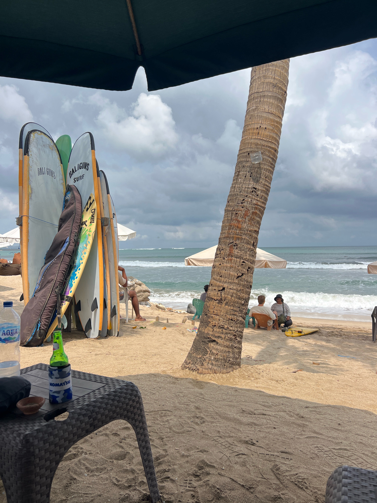 This image depicts a beach scene in Kuta, Indonesia, under a cloudy sky. In the foreground, a woven table holds a plastic bottle of AQUA water, a green bottle with a blue and white cooler sleeve featuring the word "KOMATSU", and a small brown bowl. Beyond the table, several surfboards are propped upright, some with the brand "BALI GUNS SURF" visible. A black surfboard bag leans against them. In the mid-ground, the sandy beach meets the ocean, with waves rolling in. Two people are seated in chairs facing the water, and another person is seated further down the beach. A large palm tree trunk dominates the right side of the frame, casting a shadow. Beach umbrellas are visible in the background, suggesting a resort or tourist area. The overall atmosphere is relaxed and suggests a typical day at a popular Indonesian surf destination.