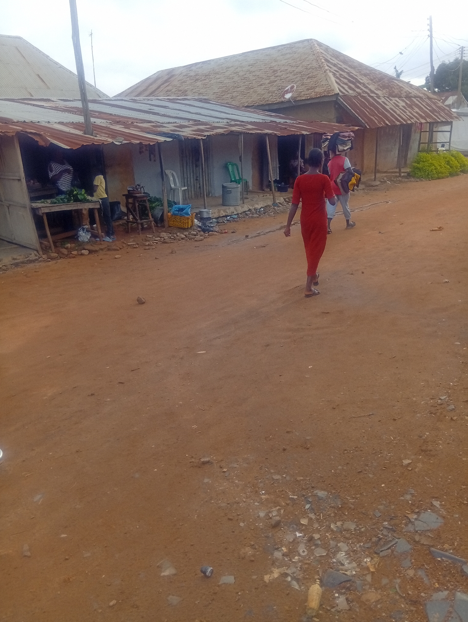 The image depicts a street scene in Yelwa, Nigeria, with a focus on a humble marketplace. In the foreground, a reddish-brown dirt road stretches into the distance. To the left, a row of market stalls with corrugated tin roofs are visible. One stall has a wooden table displaying what appear to be green produce, possibly vegetables. A person in a striped shirt stands behind the table, interacting with a vendor in a yellow shirt. Other market items, including plastic containers and cooking pots, are scattered around the stalls.
In the middle ground, a young person wearing a bright red dress walks away from the camera, heading towards the market stalls. Further in the background, two other individuals are seen walking in the same direction. One of them is carrying a bundle of clothes on their head. The buildings in the background also feature corrugated tin roofs, indicative of local architecture. The overall environment appears to be a dusty, rural or semi-urban setting. The sky is overcast.