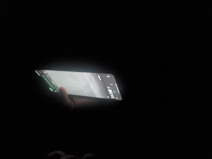 In a dark setting, a hand holds a lit smartphone at an angle. The screen displays what appears to be a web page with dense text or data. The illumination from the screen provides the only visible light, partially revealing the hand and suggesting a surrounding environment of deep darkness.