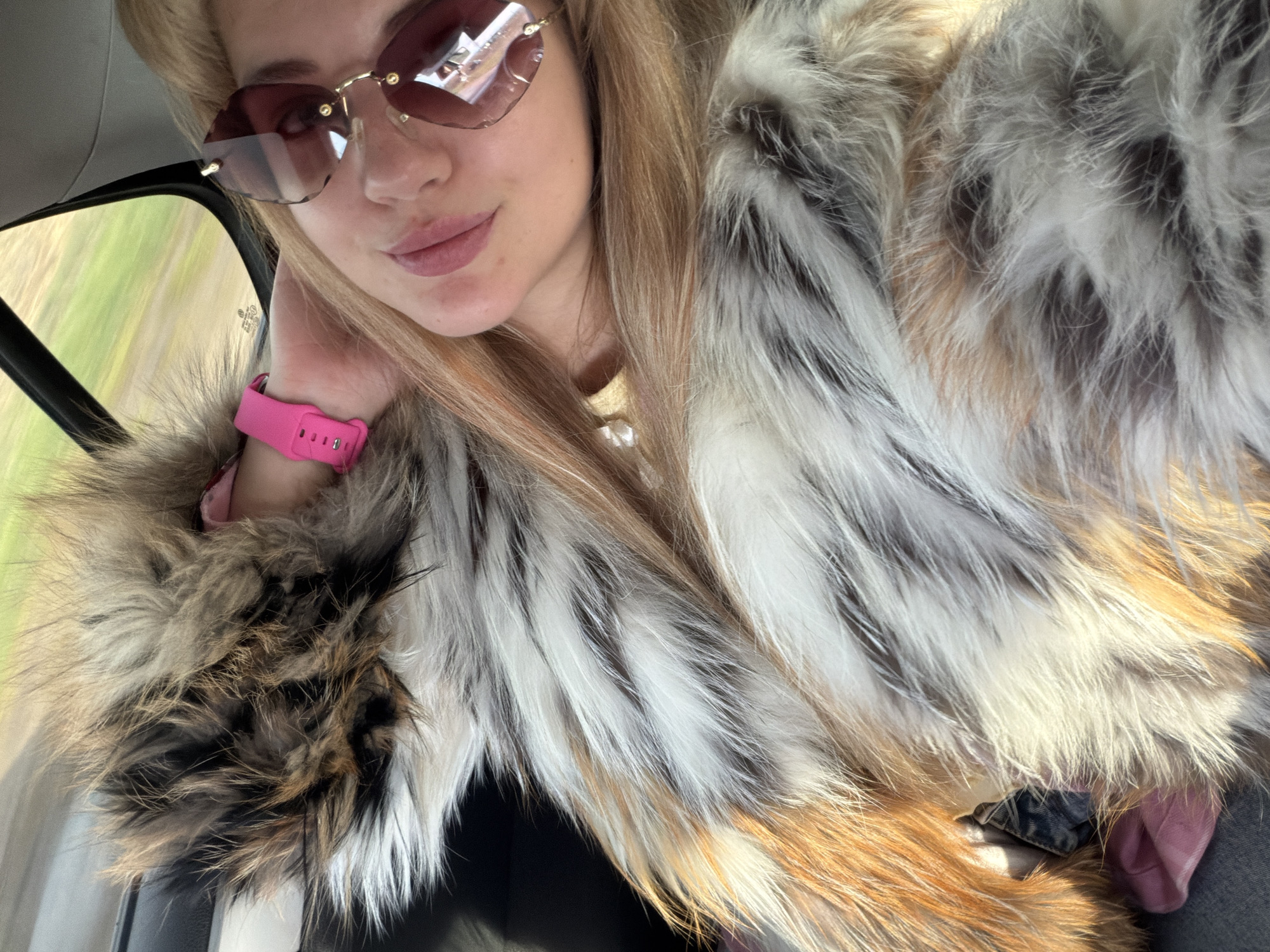 A close-up image shows a young woman seated inside a vehicle, likely a car, in Az'mushkino, Russia. She is looking towards the camera, possibly taking a selfie, with a relaxed expression and a subtle smile. She wears large, rimless sunglasses with brown-tinted lenses and gold hardware. Her blonde hair frames her face and flows over her shoulders.
She is dressed in a distinctive fur coat or collar, characterized by a varied pattern of white, brown, and black fur. A bright pink wristband, possibly a watch or fitness tracker, is visible on her left wrist, which is positioned near her chin. Through the car window on the left, a blurred green landscape indicates that the vehicle is in motion. Bright sunlight illuminates the scene, suggesting daytime and clear weather. A small, illegible sticker is present on the car window. The setting captures a moment of travel.