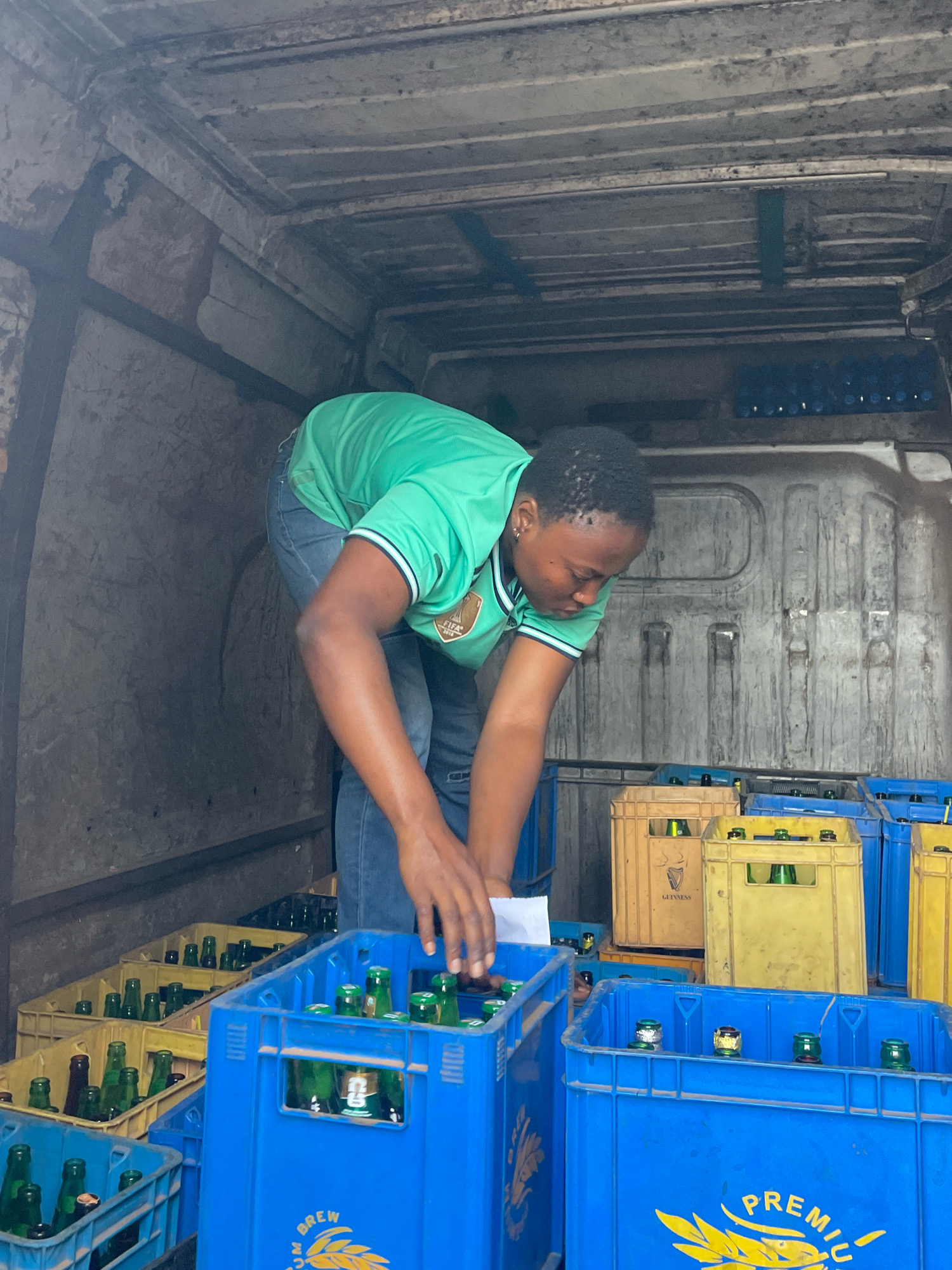 In a dimly lit interior space, possibly the back of a vehicle or storage area in Jos, Nigeria, a woman is actively engaged in organizing crates of bottled beverages. She is wearing a bright green t-shirt with white trim and a FIFA logo patch, and blue denim jeans. Her hair is short and dark, and she has earrings in her ears. The woman is bent over, her hands occupied with what appears to be a white piece of paper, suggesting she might be checking inventory or a delivery manifest.
Surrounding her are numerous crates filled with green glass bottles. The crates are of various colors, predominantly blue and yellow. Some of the yellow crates bear markings, including a recognizable "Guinness" logo. The bottles themselves are capped, and some have visible labels. The environment appears utilitarian and slightly grimy, with metal structures overhead and textured walls. The overall impression is one of logistical work, likely related to distribution or stocking of drinks.