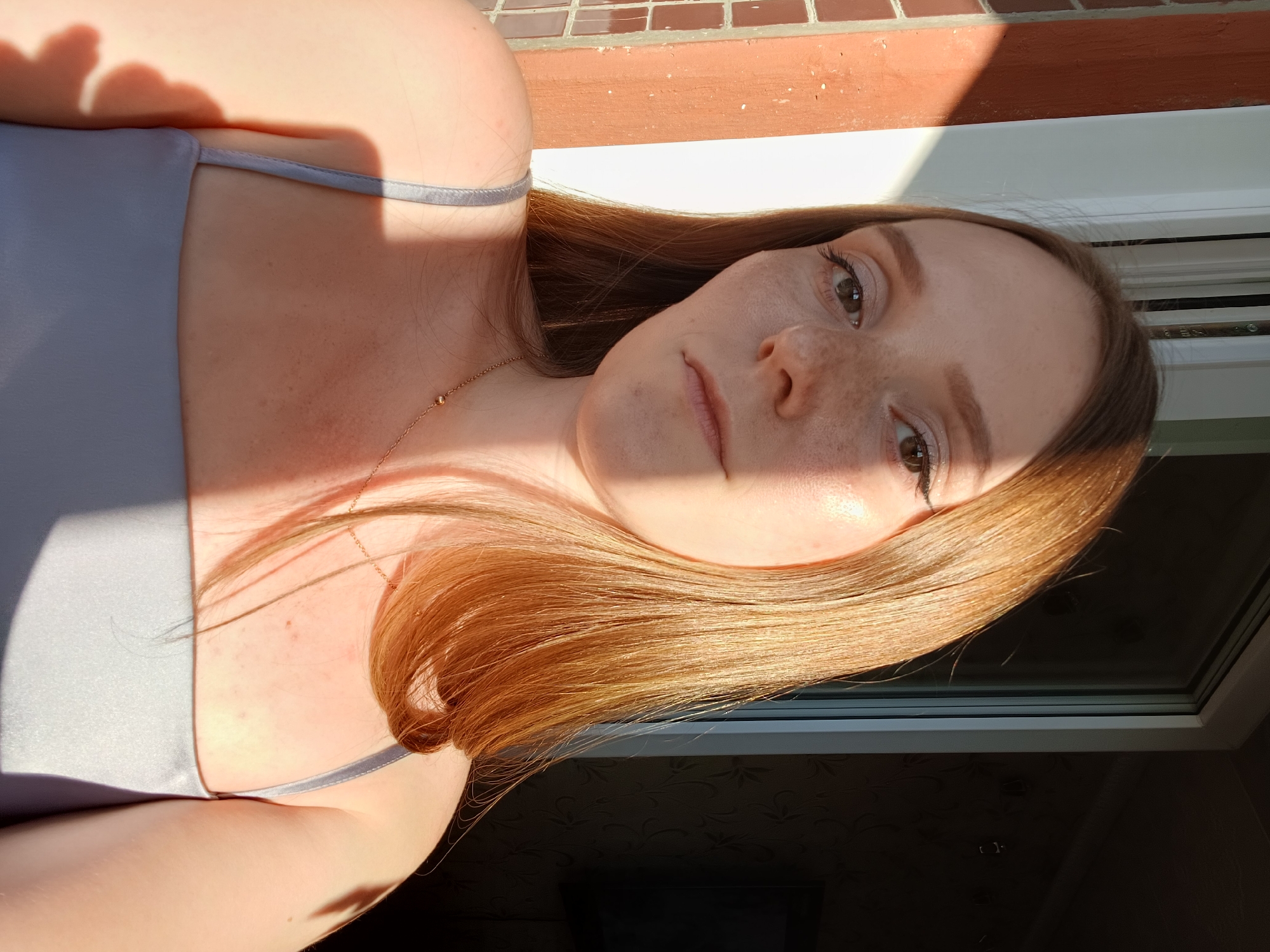 A woman, with light skin and long, auburn hair, is shown in a close-up, selfie-style shot. She is wearing a light blue, camisole-style top and a delicate gold necklace. Her face is angled towards the camera, and she has freckles scattered across her cheeks and nose. Sunlight illuminates her face, casting shadows and highlighting her features. The background appears to be outdoors, with a tiled surface and a brick wall visible in the upper portion of the image. A window with a patterned curtain is visible in the lower half of the frame. The lighting suggests it is daytime, likely late afternoon or early morning due to the warm, angled sunlight. The woman's expression is neutral, and the overall mood is relaxed and casual. There is no visible text in the image.