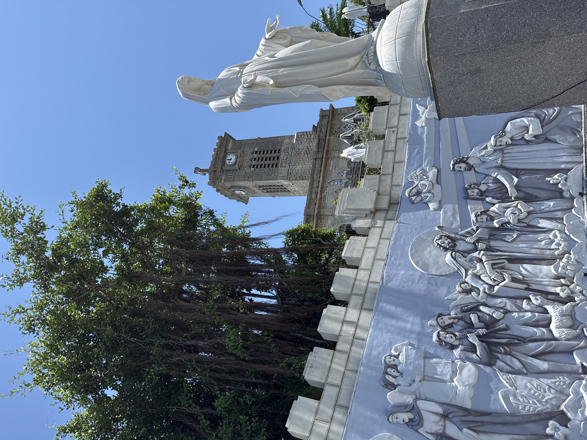 This is a low-angle, full shot of a white statue of Mary standing in front of a stone church with a clock tower. Behind the statue, a large, ancient banyan tree with hanging roots dominates the left side of the frame. In front of the church, a blue wall features a large bas-relief sculpture depicting biblical scenes with multiple figures, including angels, shepherds, and Mary holding a baby. The scene is set against a clear blue sky. The location is the Nha Trang Catholic Church in Nha Trang, Vietnam.