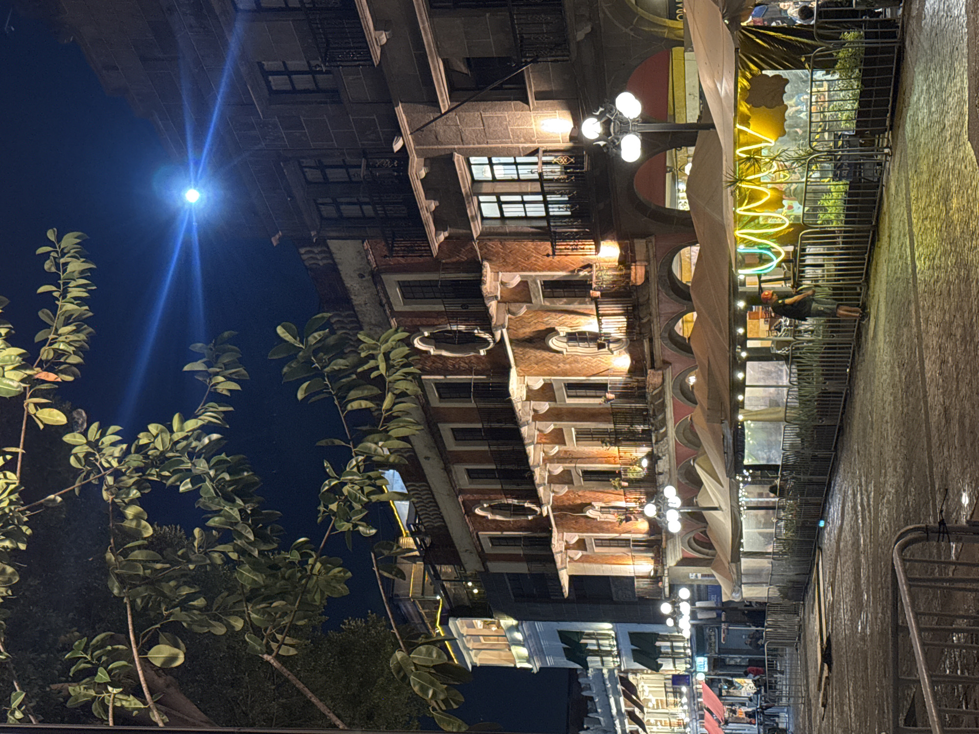At night, the image depicts a street scene in Puebla, Mexico. The buildings are made of brick and stone. Balconies are visible on several of the buildings. The road appears damp. Streetlights illuminate the street. A tree is visible to the left of the image. Security barriers line the right side of the street. Some people are visible in the distance and on the sidewalk in front of the illuminated storefronts.