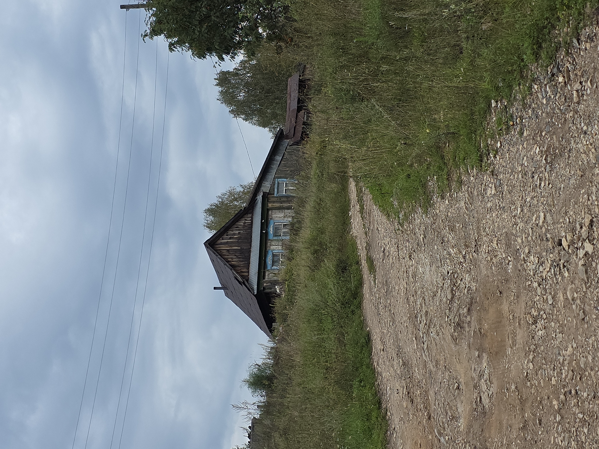 The image depicts an overcast day in Magnitka, Russia. A dirt road curves through tall, green grass. On the right side of the road, a weathered wooden house with a dark roof and blue-trimmed windows is visible. Behind the house, trees create a dense background. Power lines stretch across the sky above the scene.