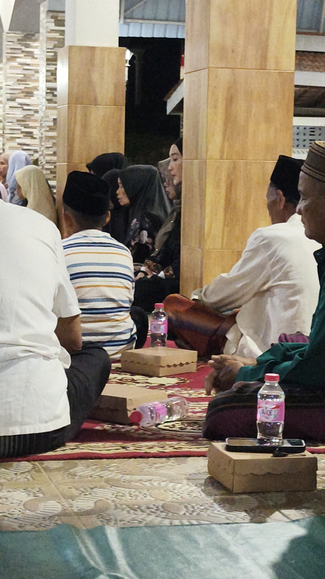 A group of people, some wearing Islamic attire, are gathered on the floor of a room. In the foreground, a person in a white shirt is partially visible from the back, seated on the floor. Next to them, a person in a striped shirt and a peci (a cap) is also seated. In the middle ground, a man in a white shirt and sarong is seated, facing away from the camera. To his right, another person in a green shirt and a peci is seated. Several cardboard boxes and two plastic water bottles are placed on a patterned rug. In the background, other individuals are seated, some wearing hijabs. The room appears to be an indoor space with wooden pillars and a textured wall. The scene suggests a gathering or religious event in Dalegan, Indonesia.